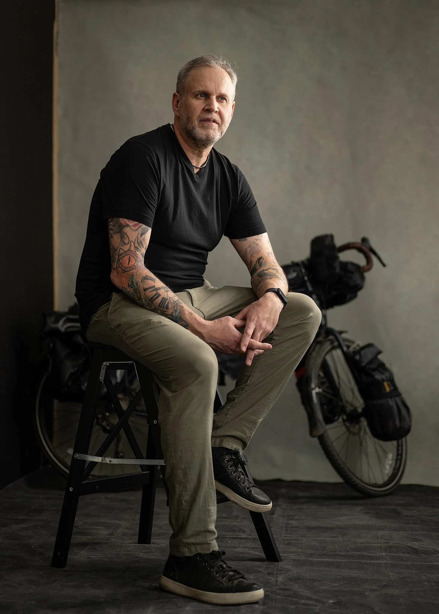 A man with tattoos on his arms, wearing a black t-shirt and khaki pants, sitting on a stool in front of a textured backdrop, with a bicycle loaded with gear in the background.