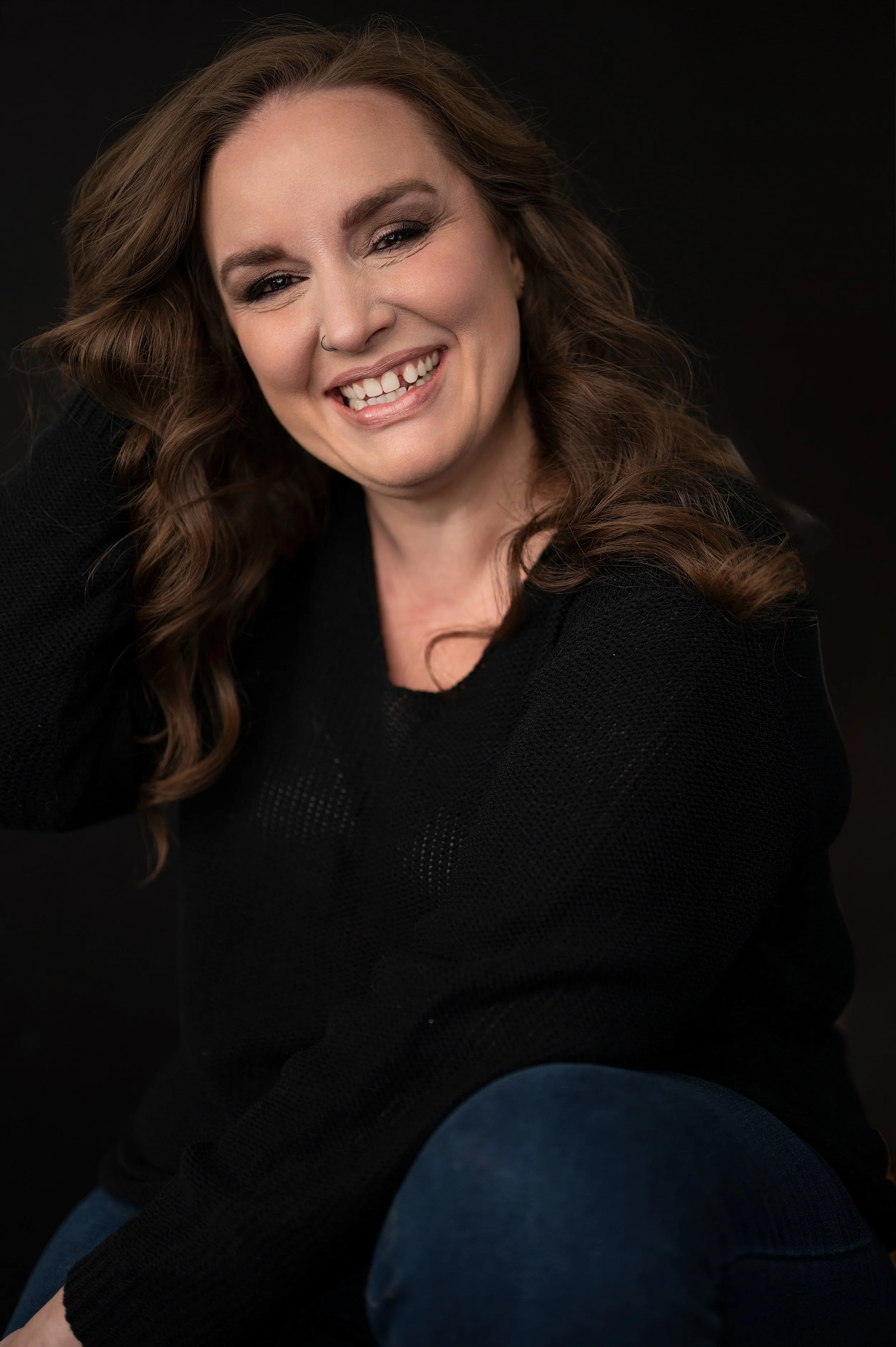 A woman with long wavy brown hair smiling, wearing a black sweater and blue jeans, against a dark background.