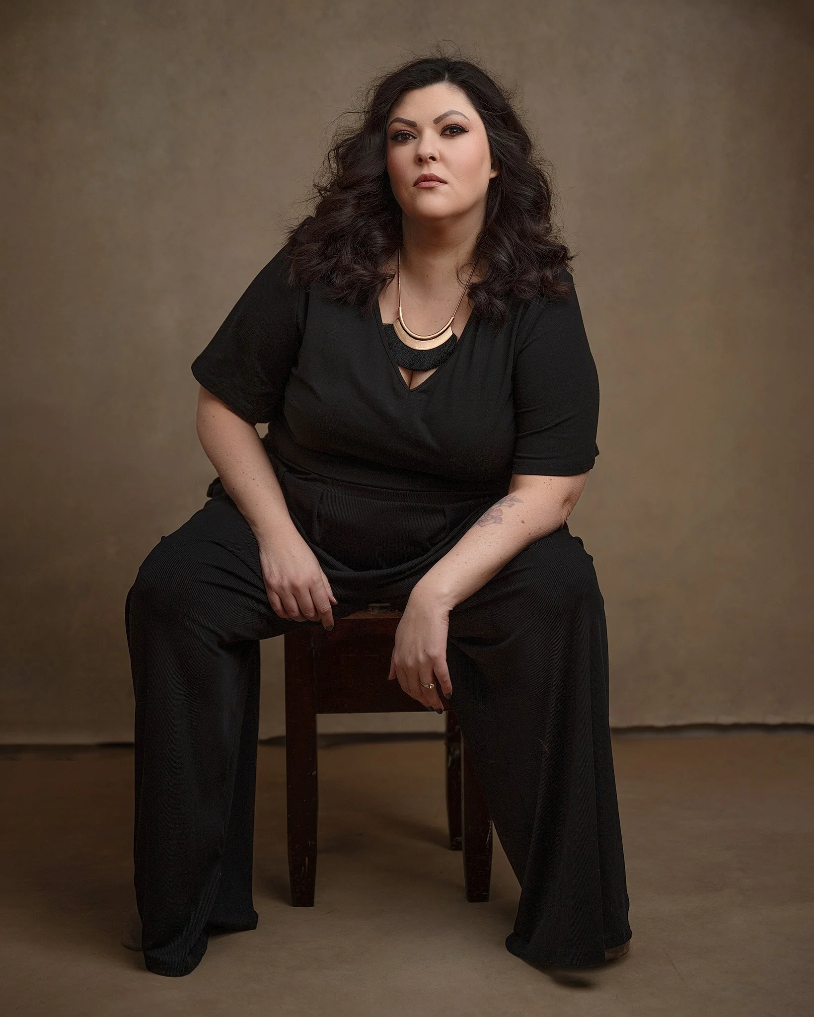 A woman with dark, curly hair and makeup wearing a black outfit with a gold necklace, sitting on a dark wooden chair against a plain beige background.