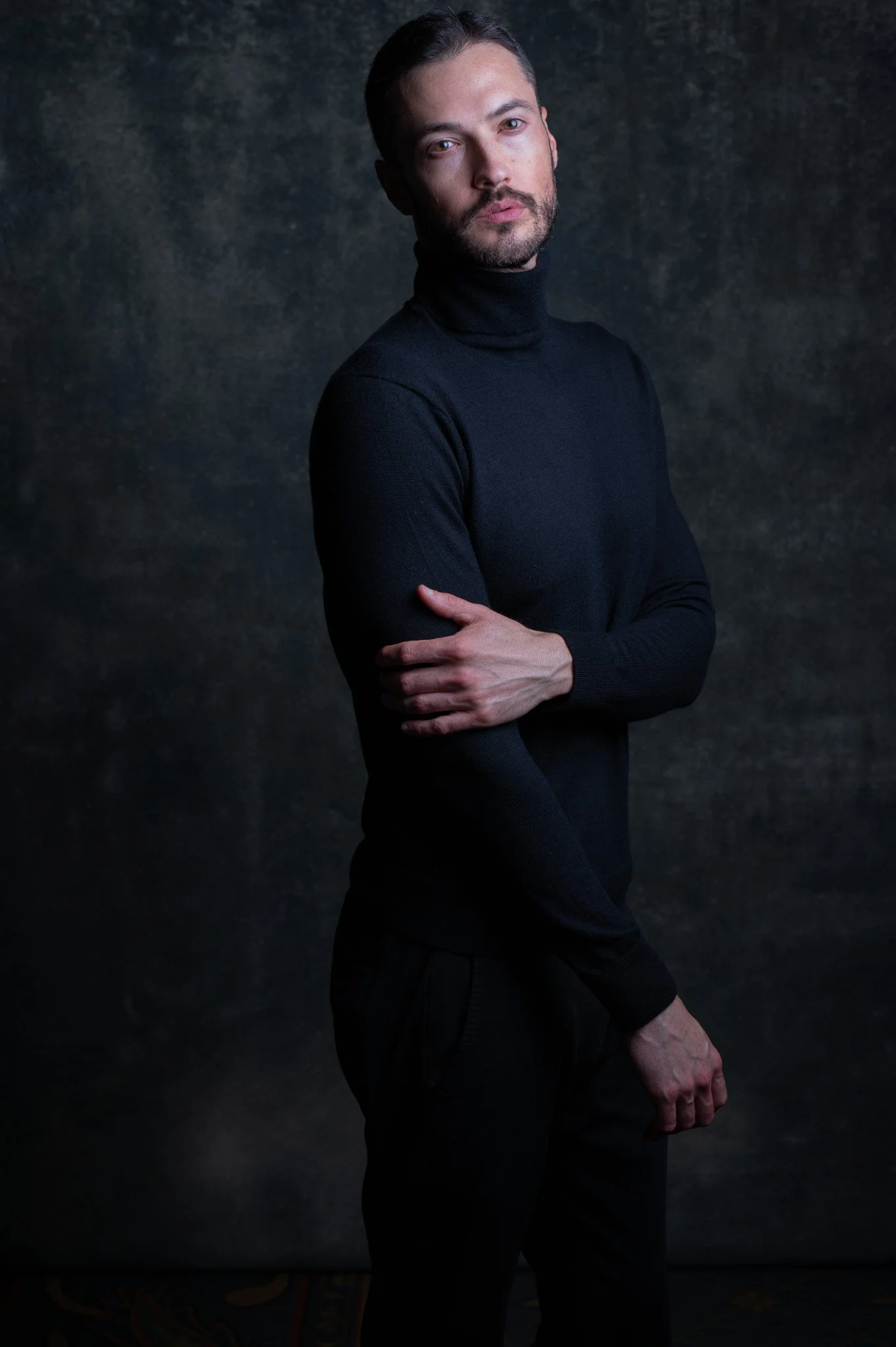 Personal brand image of a man with dark hair, beard, and light skin, wearing a black turtleneck sweater, posing against a dark textured background.