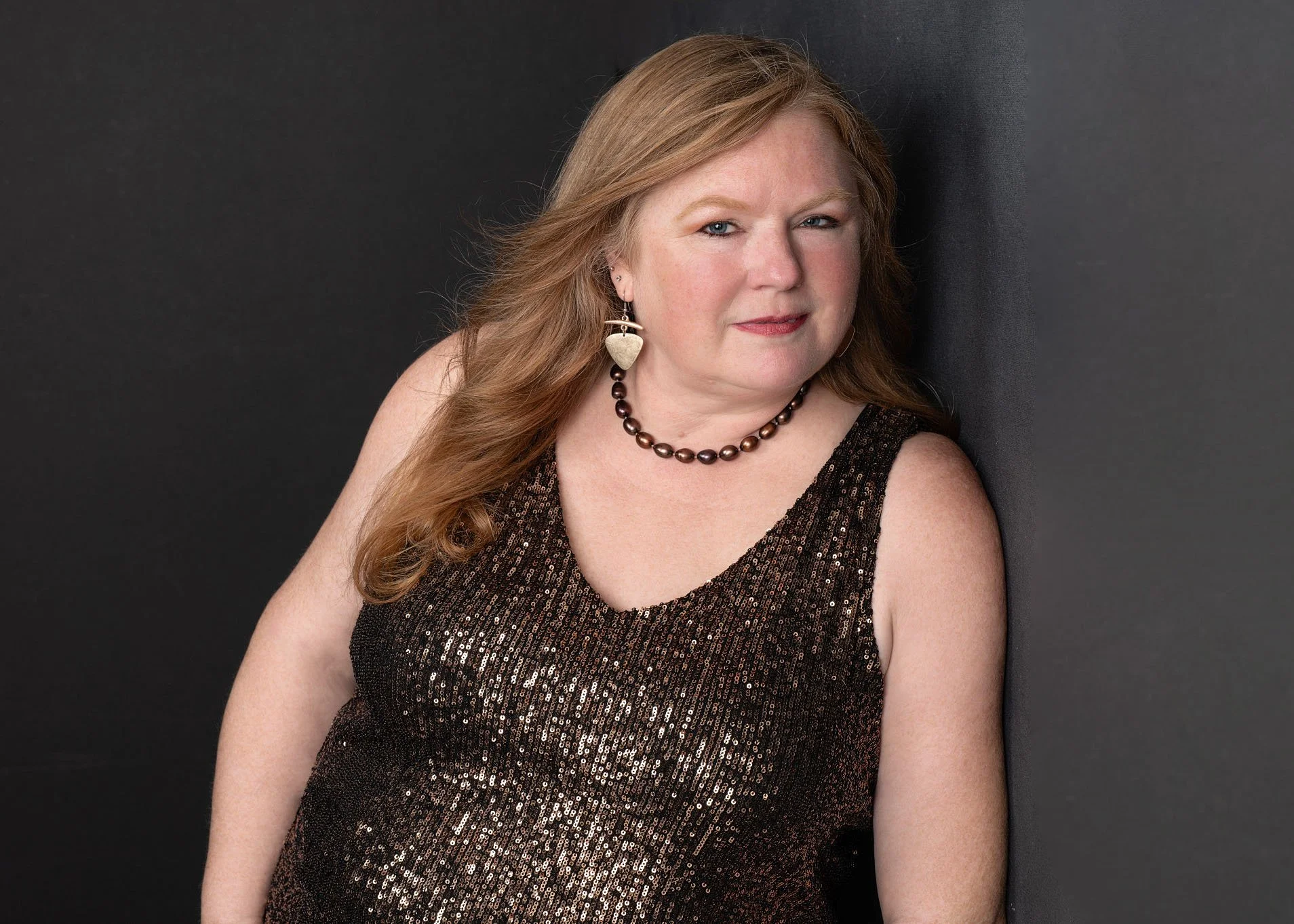 A woman with long reddish hair wearing a sparkly black sleeveless top, a pearl necklace, and earrings, posing against a dark background.