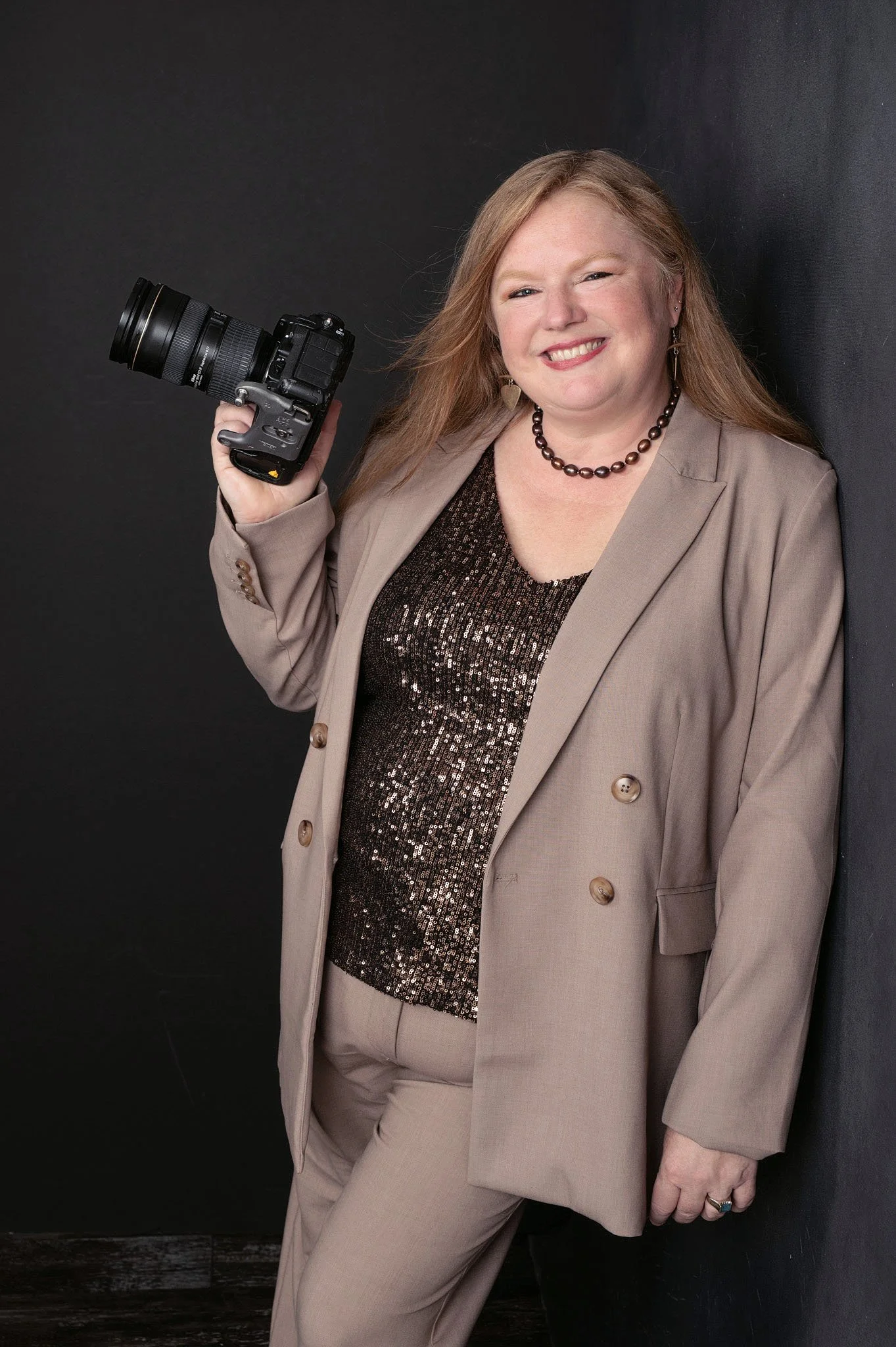 A woman with long blonde hair smiling, wearing a beige suit with a sparkling sequined top, holding a camera, standing against a dark background.