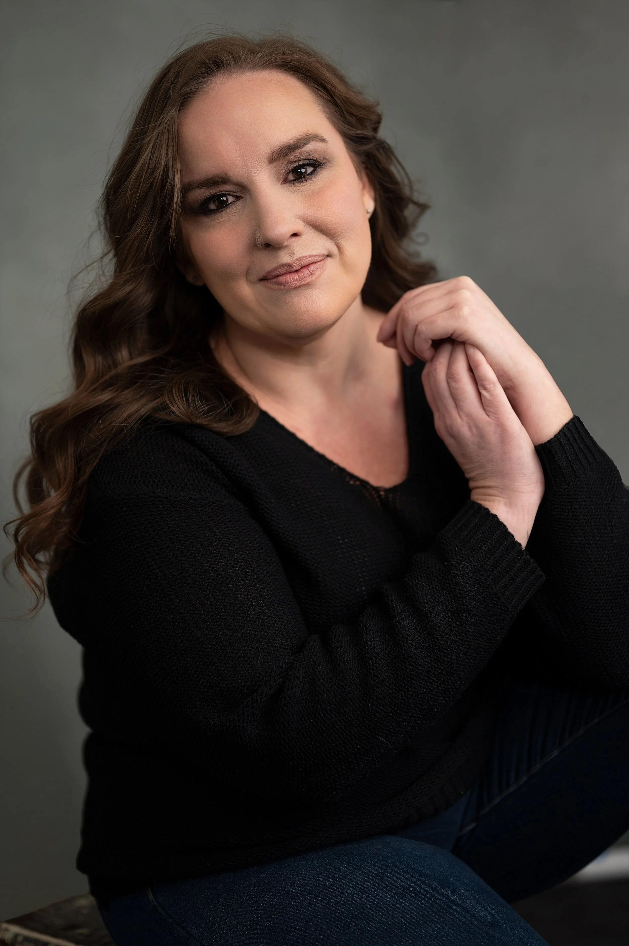 A woman with long brown hair, wearing a black sweater, sitting with her hands clasped near her face, smiling softly at the camera against a plain gray background.