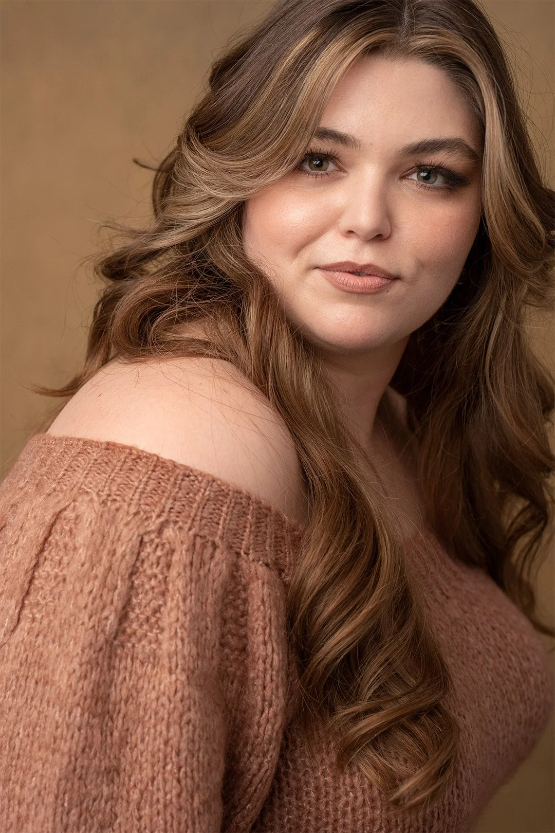 A woman with long, wavy brown hair wearing an off-the-shoulder brown sweater, looking at the camera with a slight smile, against a plain beige background.