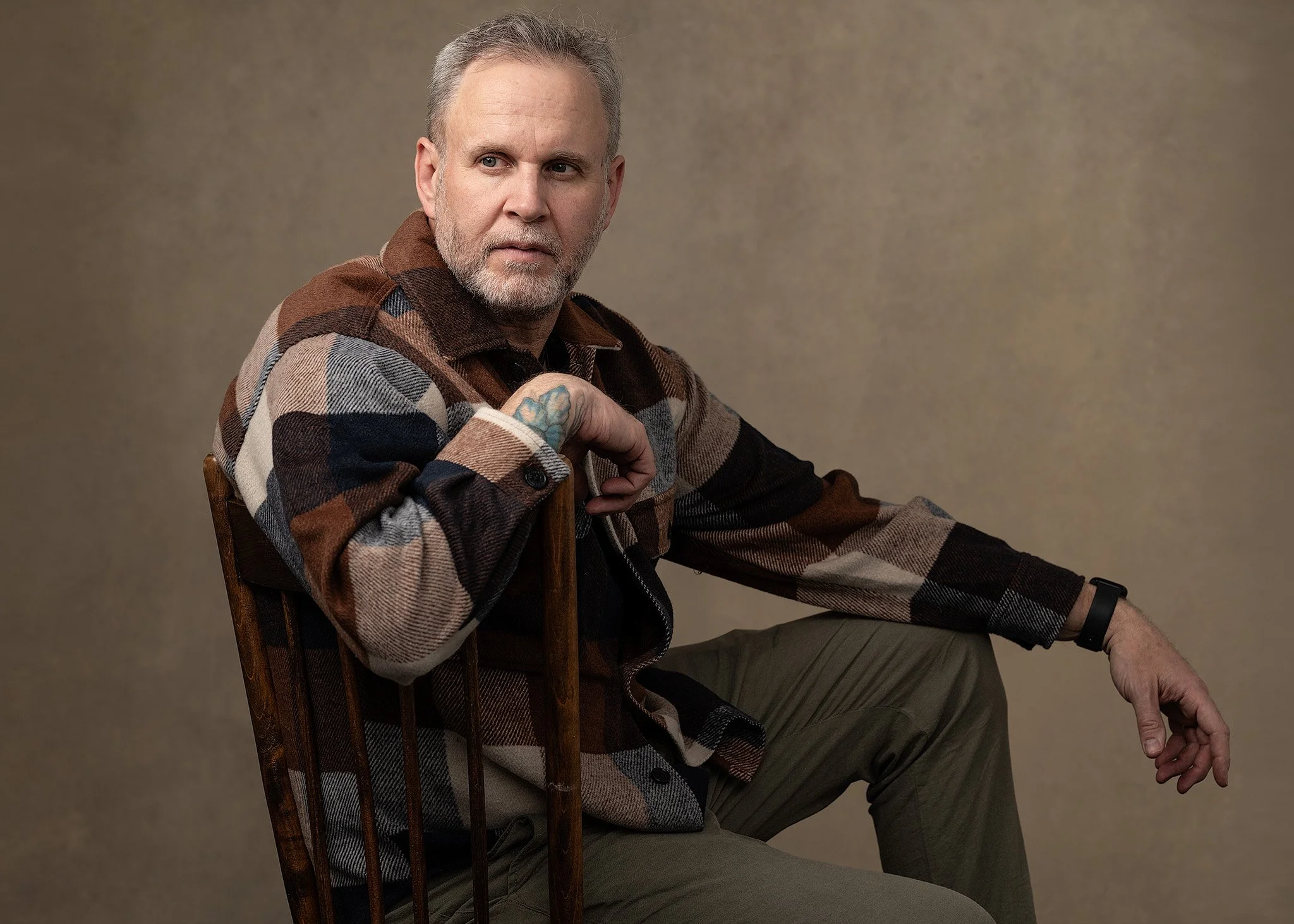 A middle-aged man with gray hair and a beard, dressed in a multicolored plaid shirt and olive green pants, sitting on a wooden chair against a plain beige background.