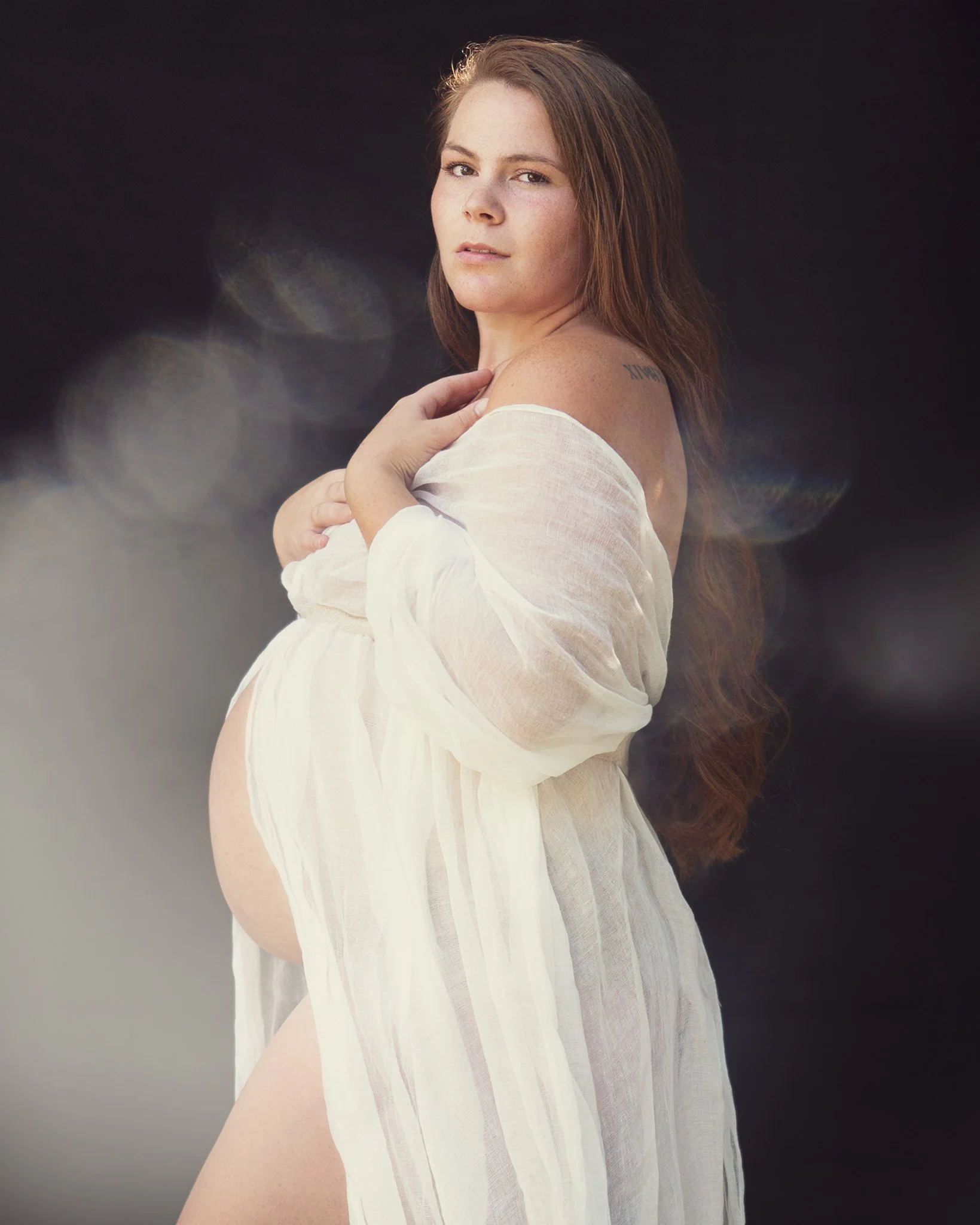 Pregnant woman wrapped in flowy gauze fabric with whimsical light flare across photo, portrait by Red Deer photographer at Carmen R Photographer photo studio.
