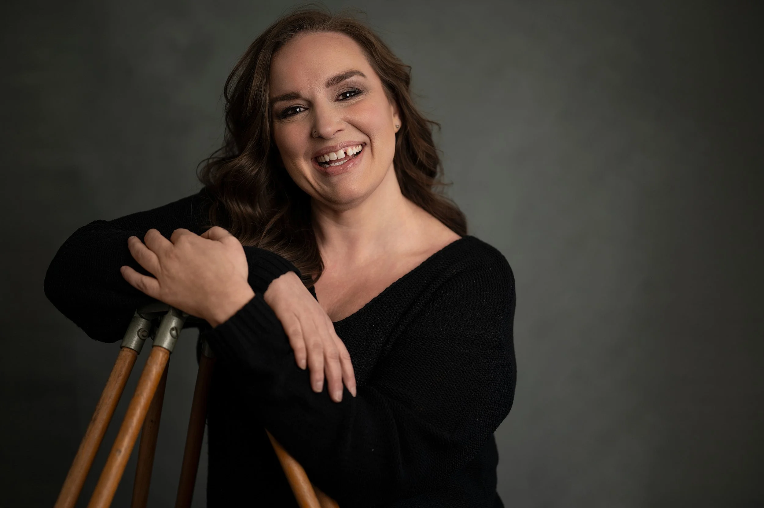A woman with wavy brown hair smiling and sticking out her tongue, leaning on wooden crutches, wearing a black sweater, against a gray background in this professional brand photograph.
