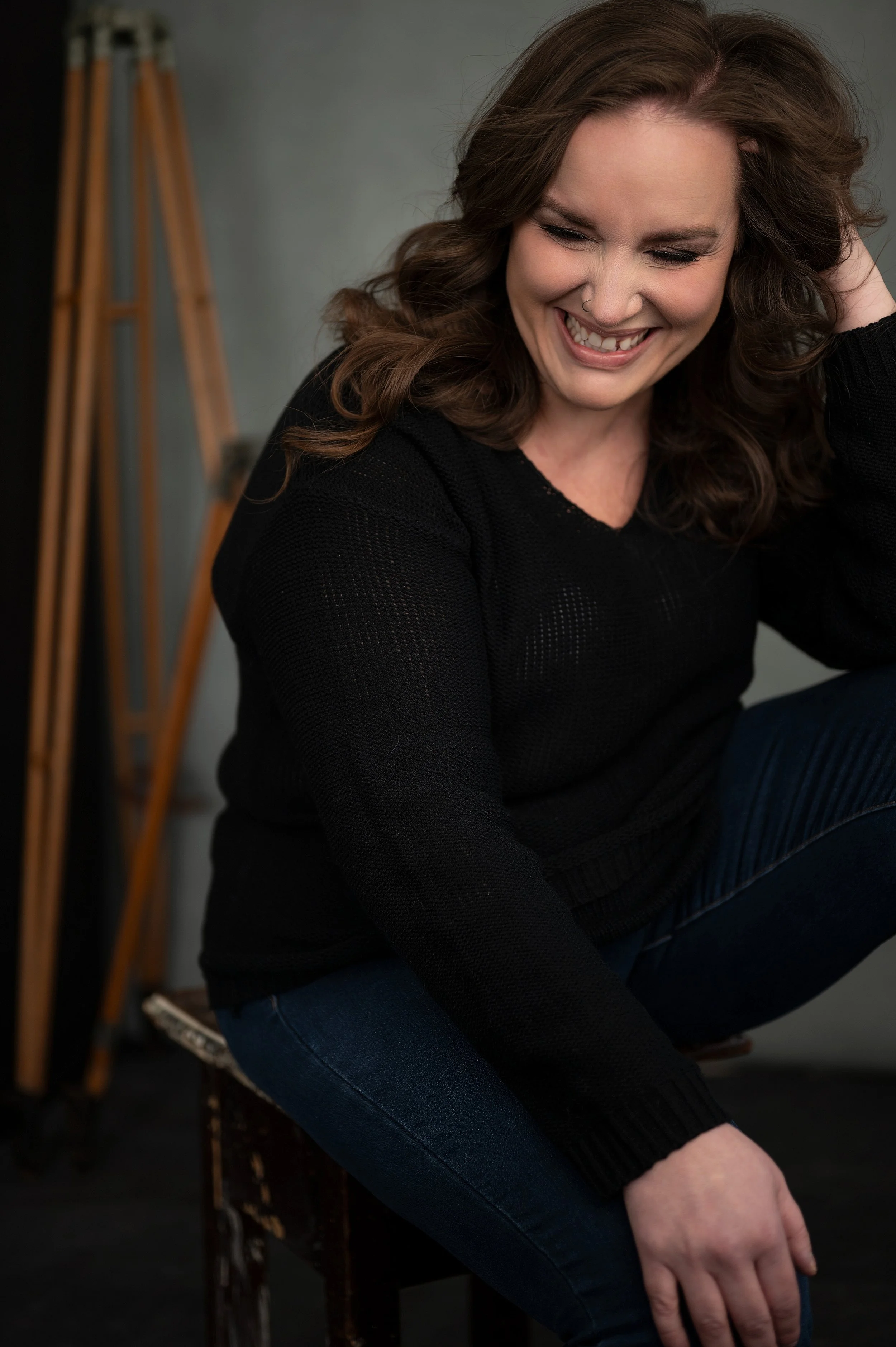 A woman with brown hair smiling and looking down, sitting on a stool in a studio, wearing a black sweater and dark jeans.