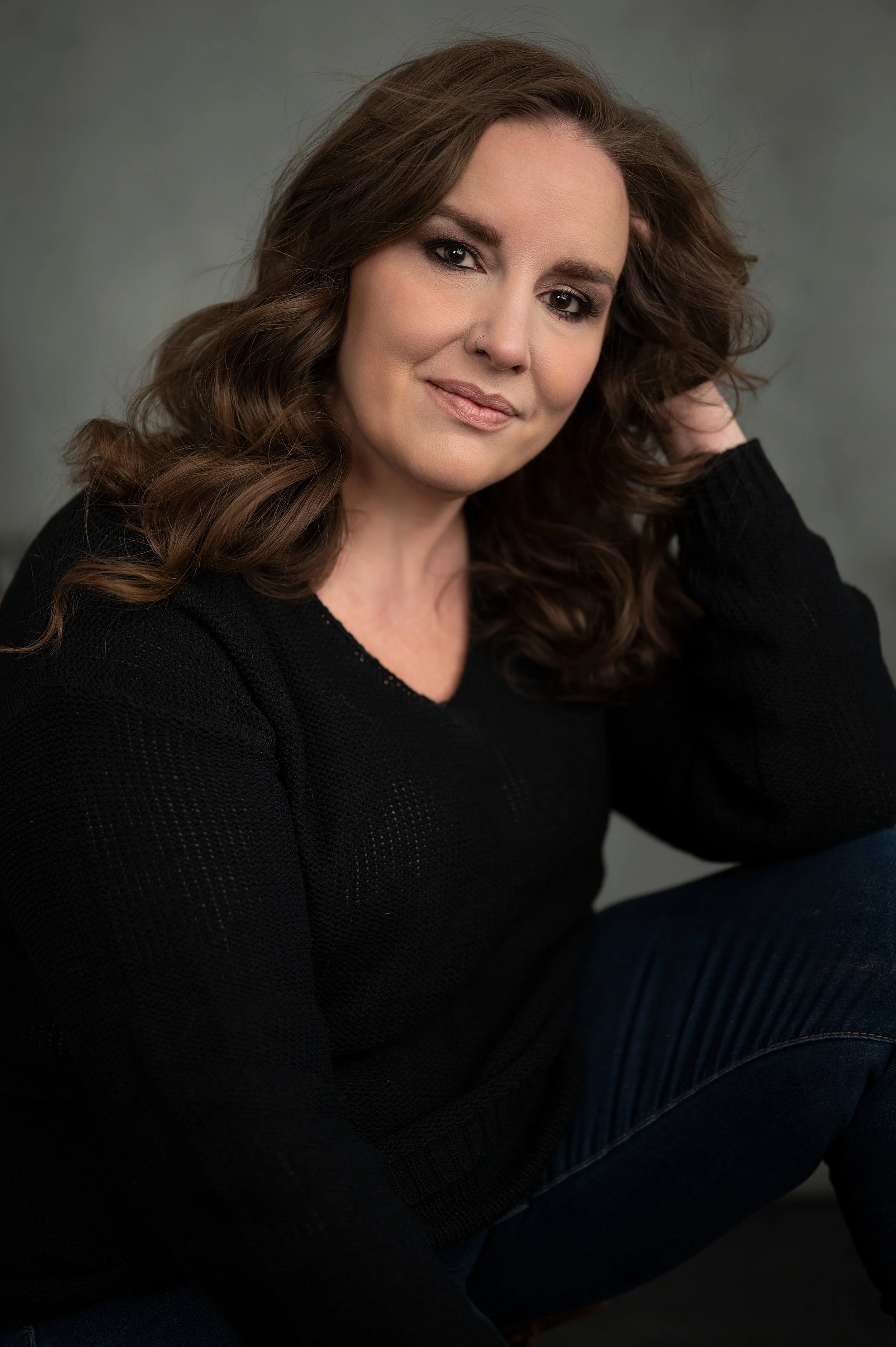 A woman with wavy brown hair wearing a black sweater, sitting against a gray background, looking at the camera with a slight smile.