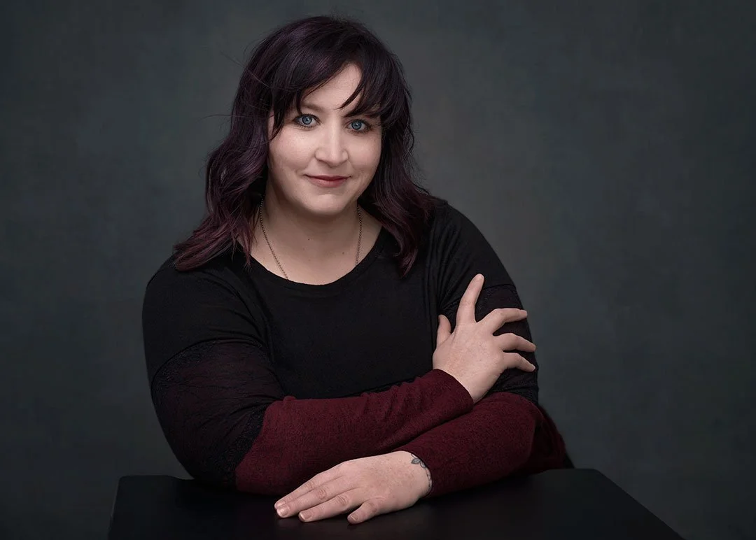 Professional branding photo of a woman with dark purple hair, light skin, wearing a black top with maroon sleeves, sitting at a black table against a dark background.