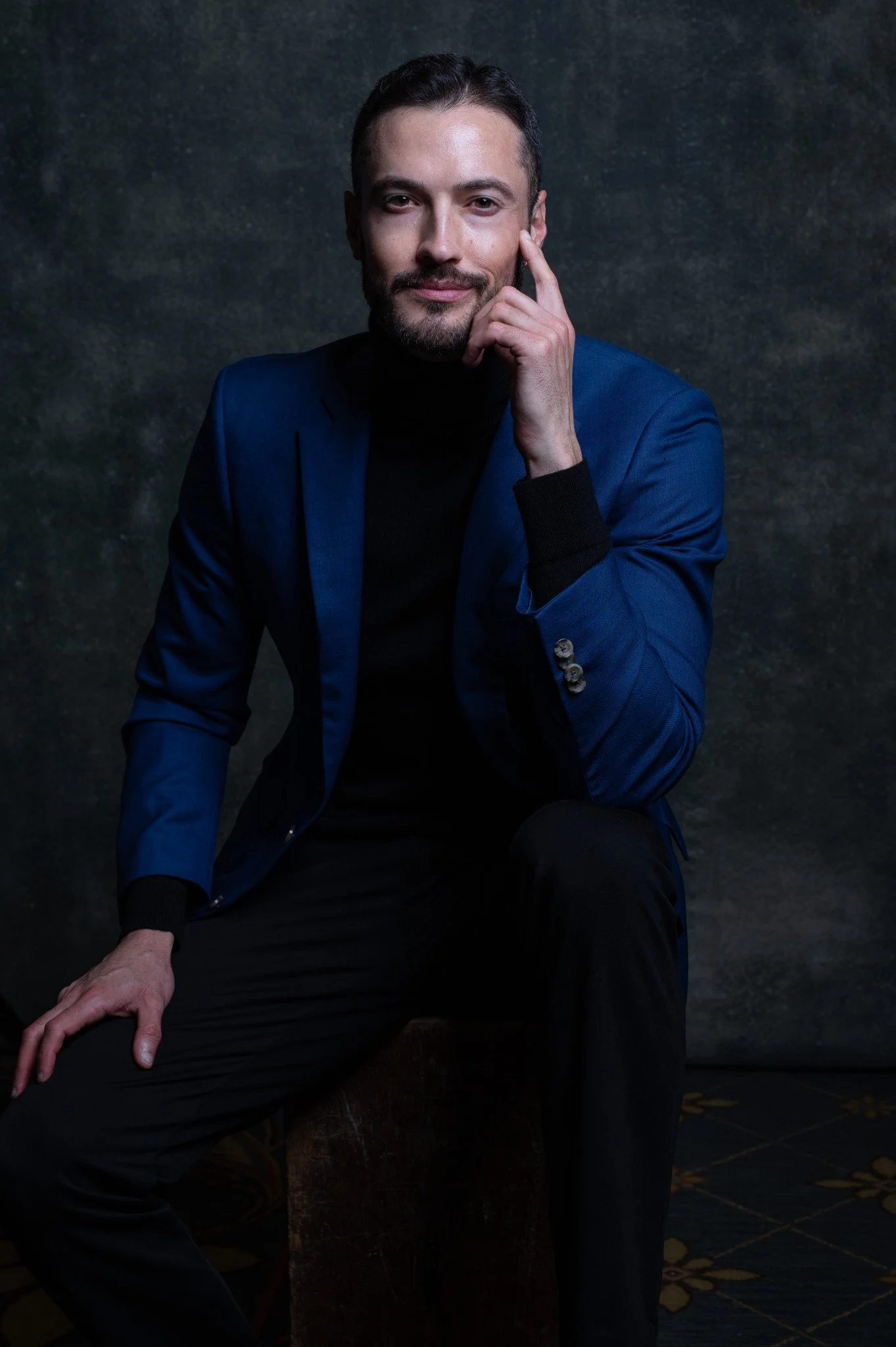 A man with dark hair, beard, and mustache sitting on a wooden box against a dark textured background. He wears a blue blazer over a black turtleneck, with black pants, and has a confident expression.