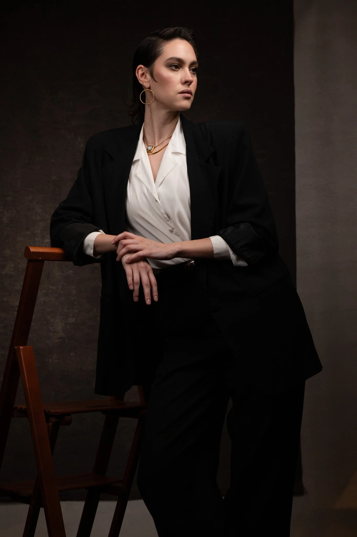 A woman with short dark hair dressed in a black blazer, white blouse, and black pants, leaning on a wooden chair, looking to the side against a dark and neutral background.