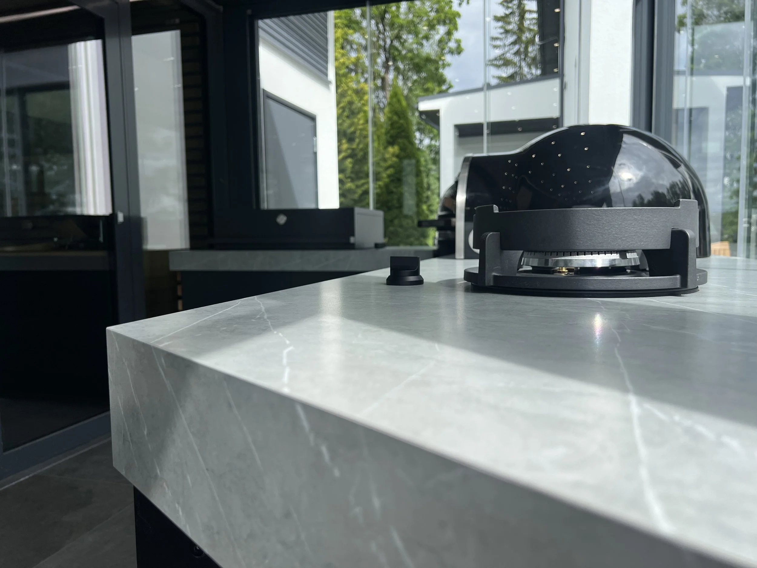 Mustapohjainen kaasuliesi keittiössä, taustalla suuret ikkunat ja vihreää ulkona.