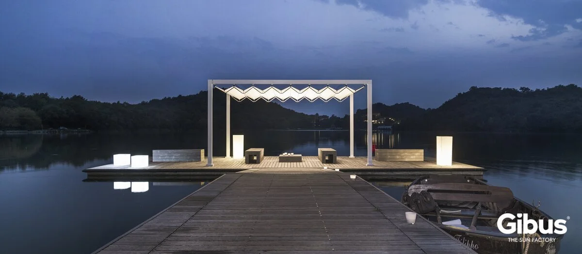 Tukeva laituri, jossa on valaistu kioskityylinen katos, istuimia ja lamppuja, ympärillä vettä, taustalla puita ja mäkiä, iltavalaistuksessa.