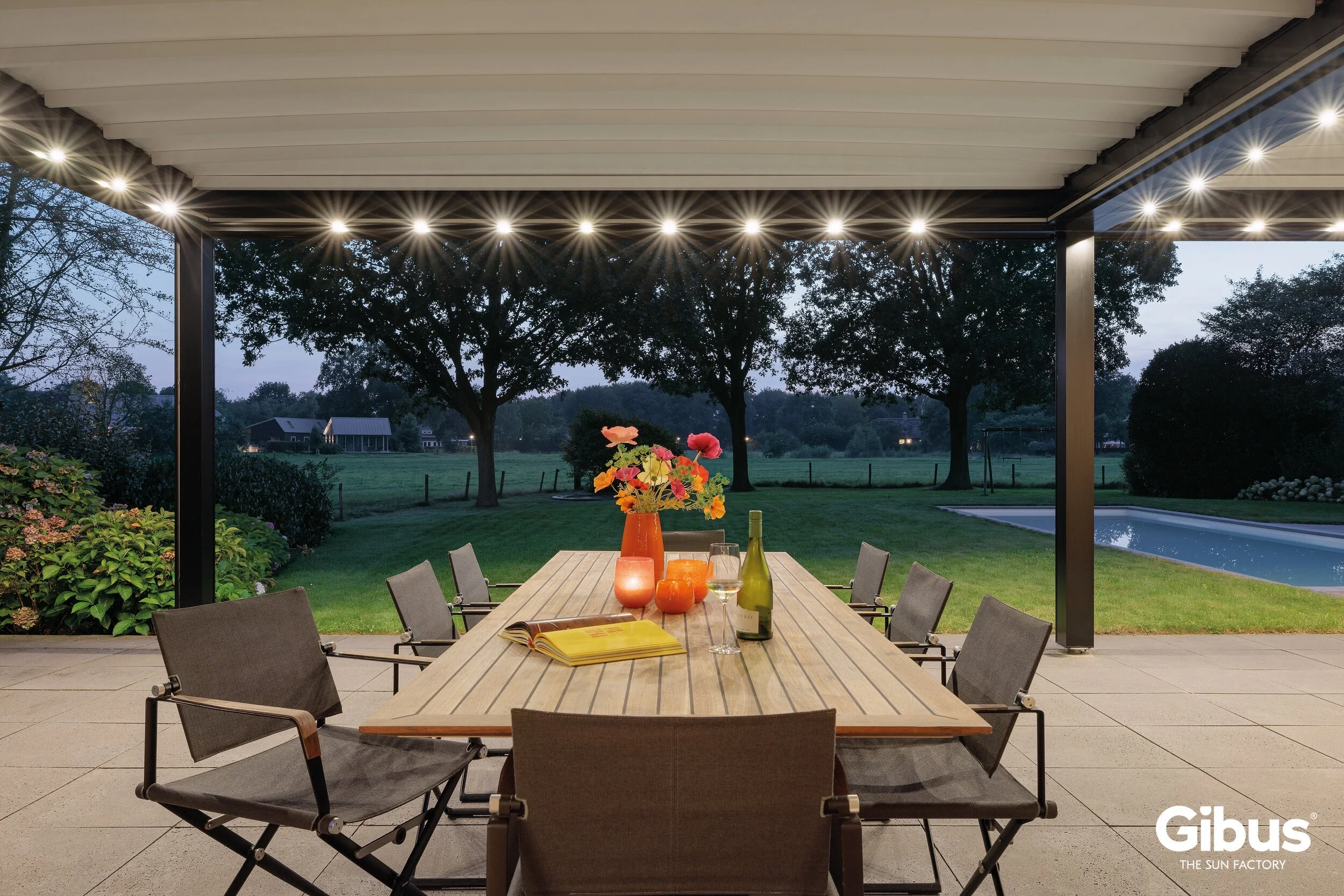 Terassi näkyy iltavalaistuksessa, pöydällä on kukkavaza, kynttilöitä, viini ja auki oleva kirja. Taustalla puut ja nurmikko, uima-allas ja maalaistaloja.