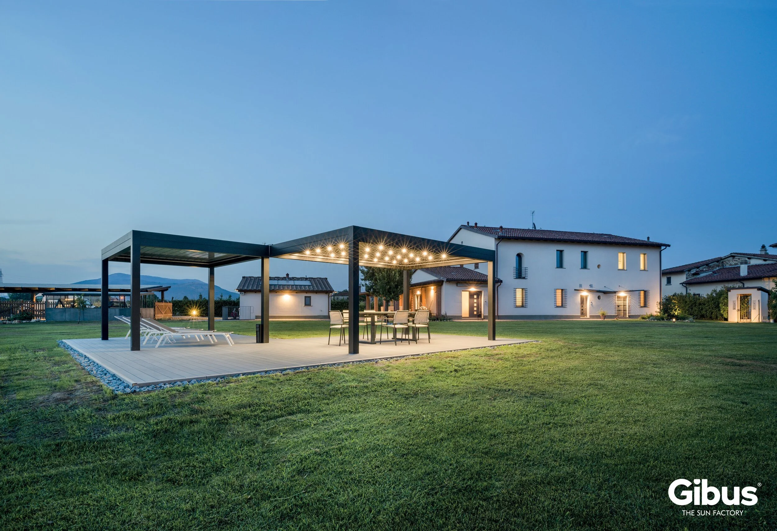 Kuva modernin terassin ja ulkotilan valaistuksesta pimeässä illassa. Terassilla on pöytä ja tuoleja, ja katos valonauhalla. Taustalla on taloja ja vuoristoa. Gibus logo alhaalla oikealla.