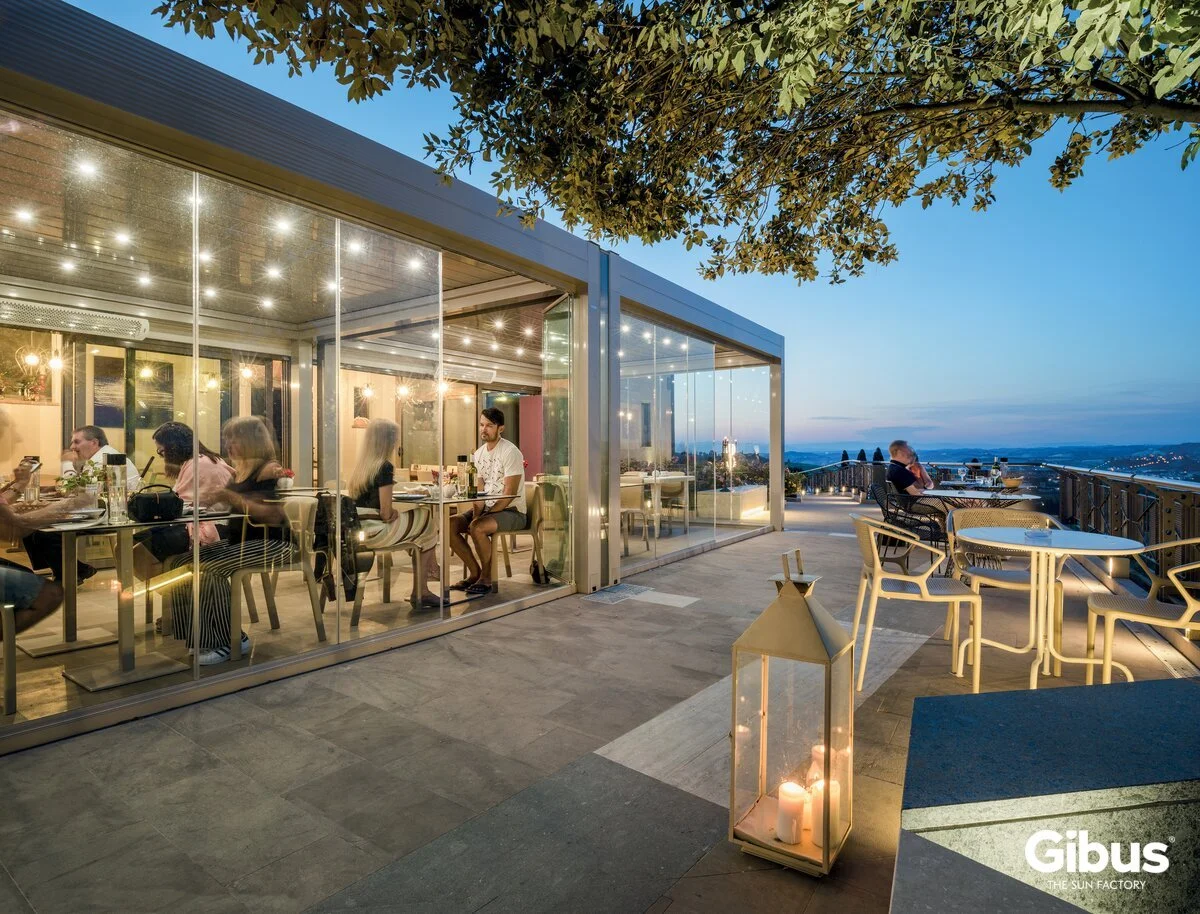 Ihmiset istuvat ravintolassa lasiseinässä sisällä, ulkona on pöytä ja tuoleja, kynttelikkö ja kaupunkinäkymä taustalla auringonlaskun aikaan.