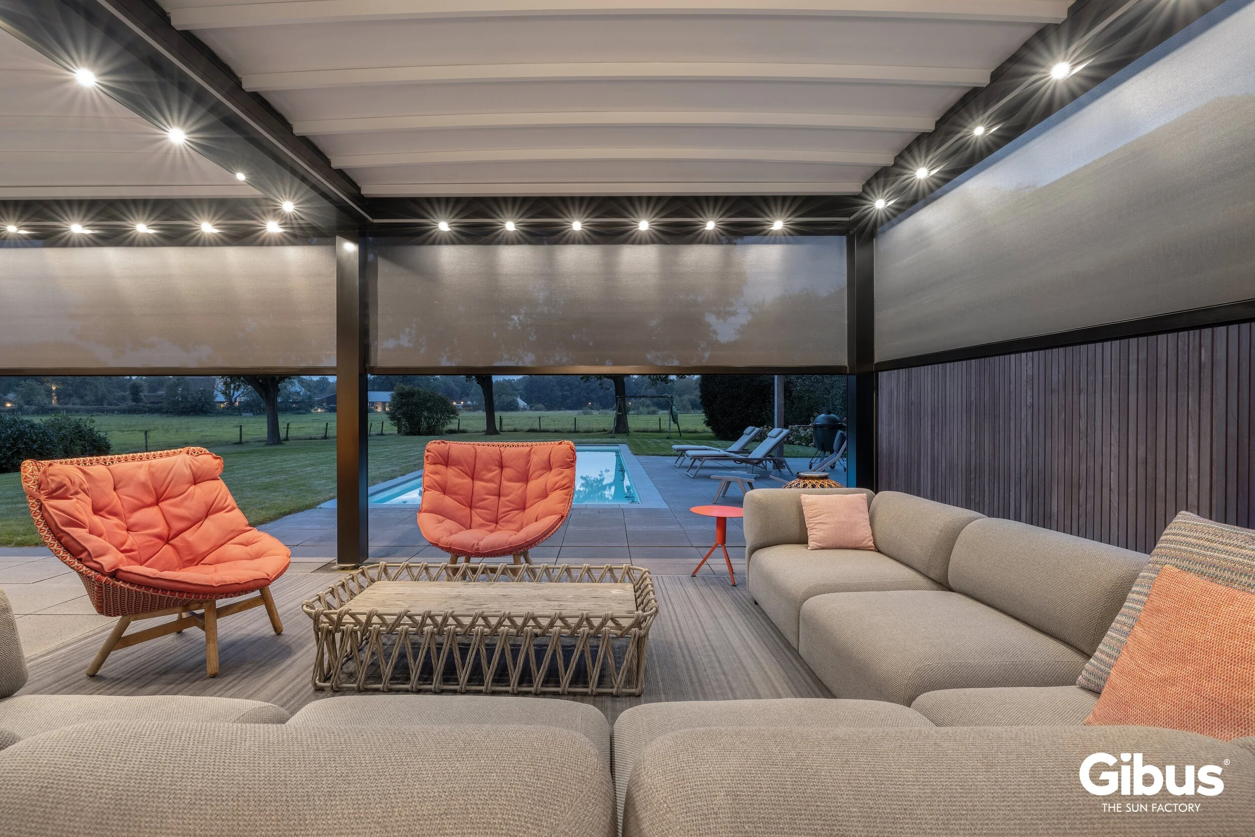 Sisätiloista terassille, jossa on suuri U-muotoinen harmaa sohva ja kaksi oranssia nojatuolia. Takana näkyy uima-allas ja ulkona vihreää nurmikkoa ja puita. Sisätiloissa on kattovalaisimia ja seinä rävipanelointia.