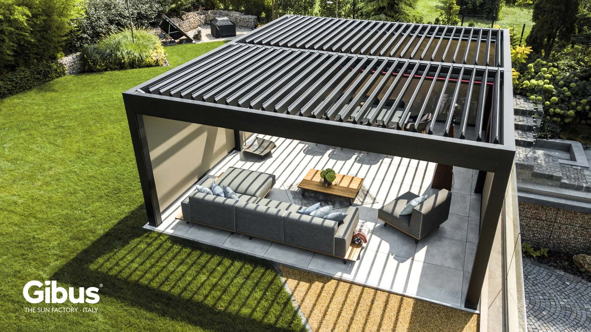 Kato: moderni ulkovarasto, jonka katto on varustettu varjostavilla rimoilla, ja jonka alla on oleskelualue, jossa on sohva, tuoleja ja pöytä, ympäröi vihreä nurmikenttä ja pensaikkoryhmä.