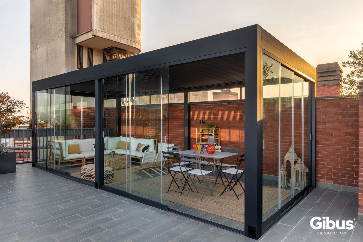 Lasitettu terassi tai oleskelutila katolla, jossa on valkoinen sohva, ruokailupöytä ja tuoleja, kynttilälyhtyjä ja koristeita, taustalla punainen tiiliseinä ja kaupunki.