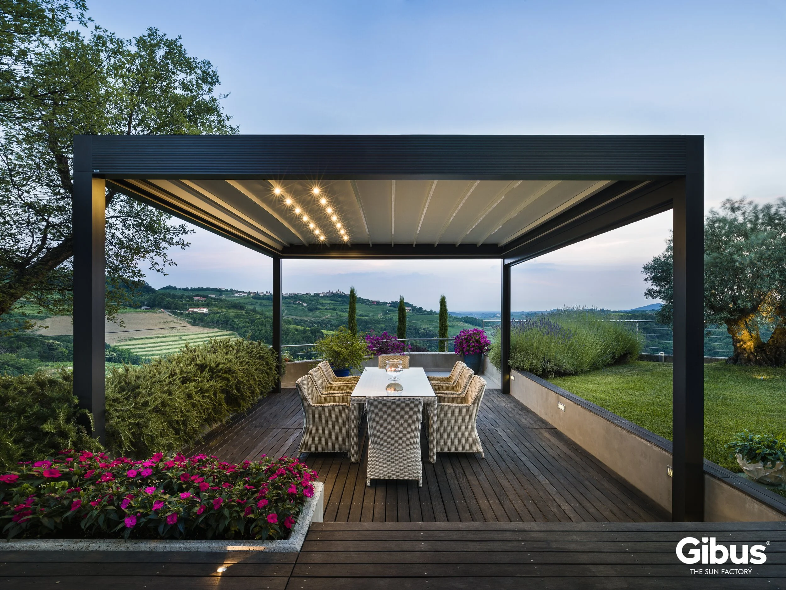 Terassi puupinnoilla ja moderneilla valaisimilla, näkymä viileälle kukkivalle pihalle rinnalla ja kaukaisiin vuoristomaisemiin iltavalaistuksen kera.