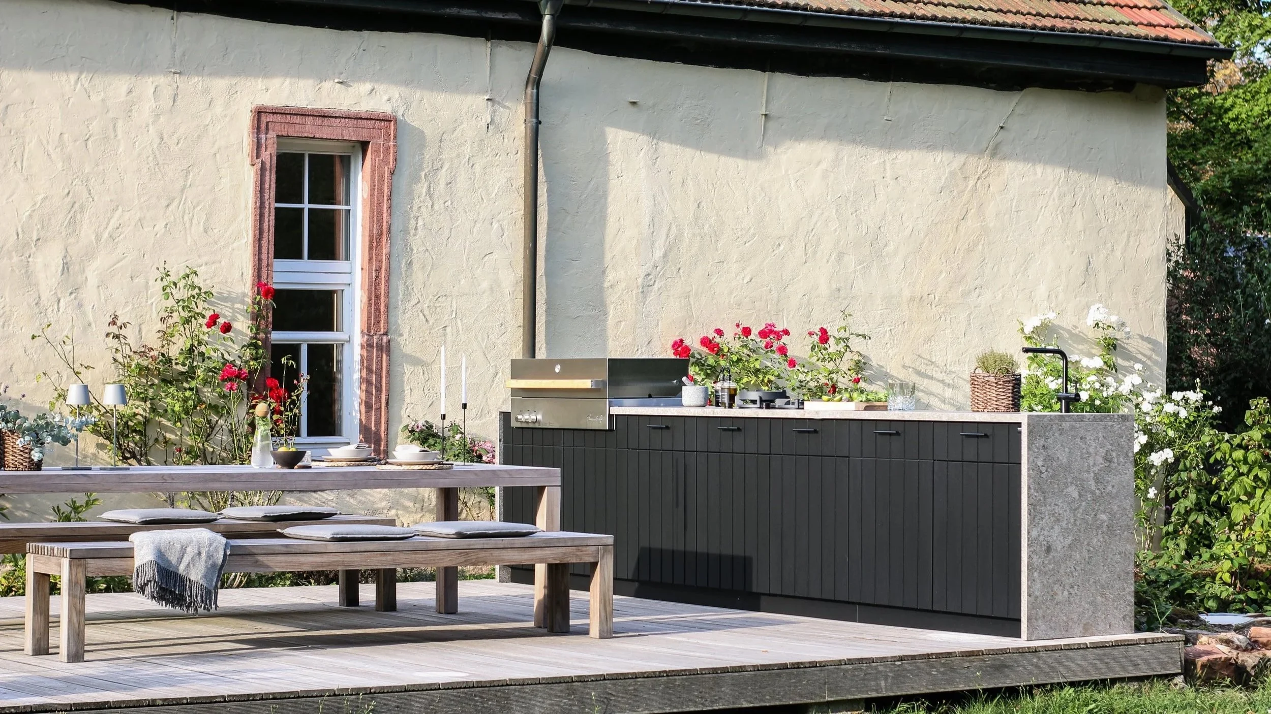 Ulkona sijaitseva keittiö ja ruokailutila, jossa on pitkät puiset pöytä ja penkki, sekä grilli ja kukkalaatikot roast emb chop, kukkia, laseja, ja kynttiläkouruja.