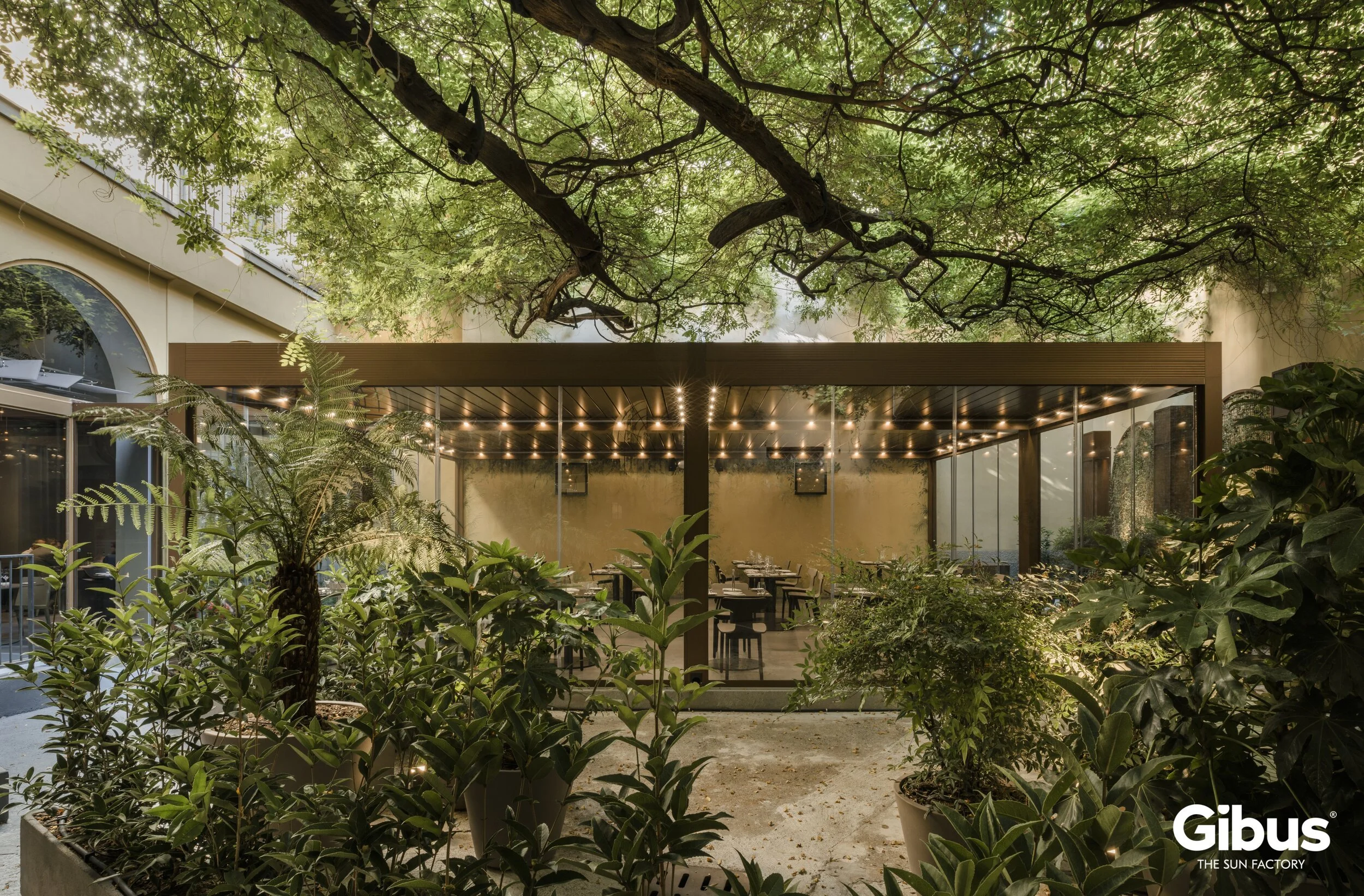 Kasvihuone, joka on rakennettu täysin lasista ja puusta, ympäröi puita ja kasveja. Sisällä näkyy pöytiä ja tuoleja, valaistus katossa.