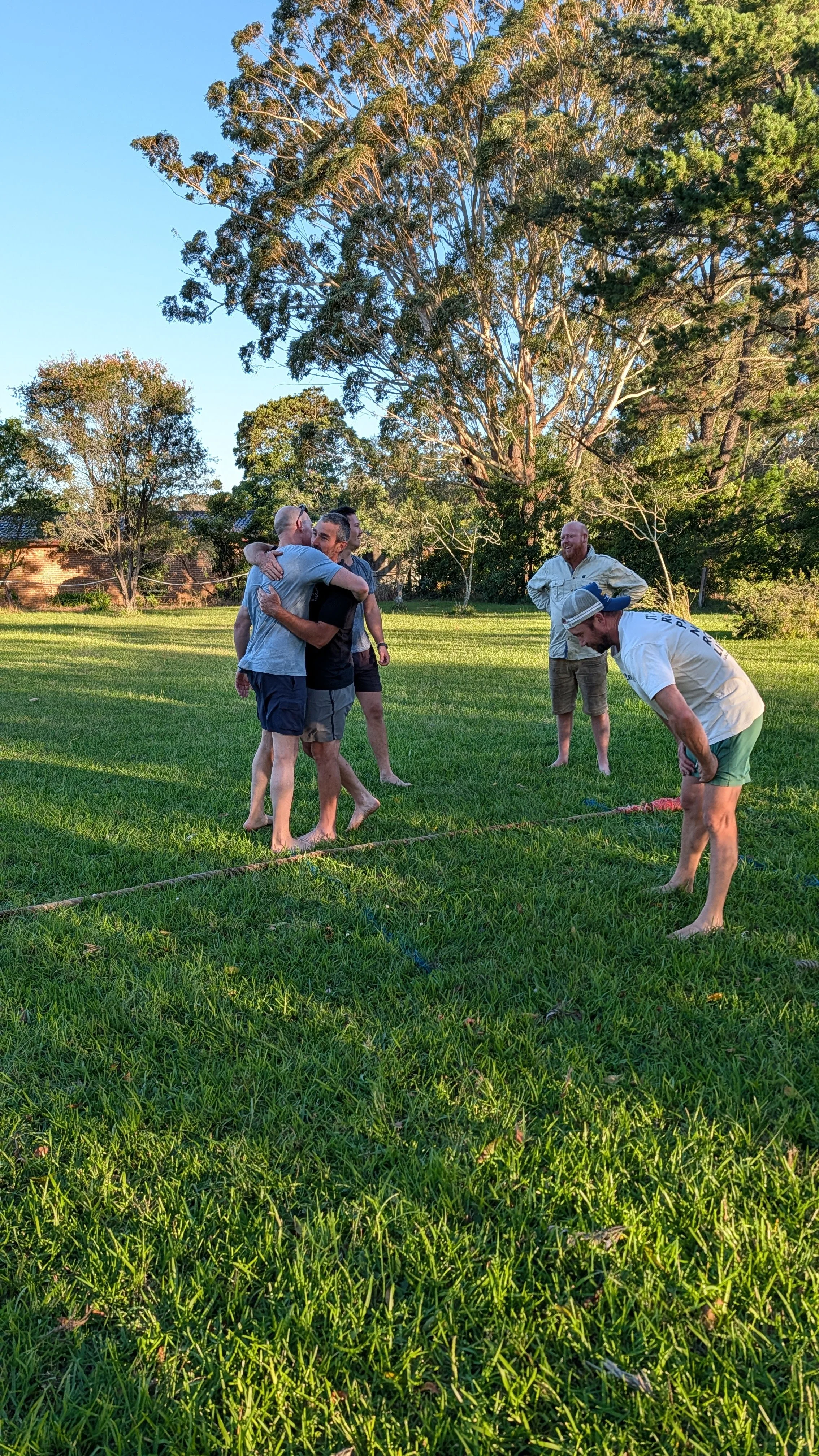 Group of five men on a grassy field, some hugging and one bending over, with trees and a house in the background, during late afternoon or early evening.