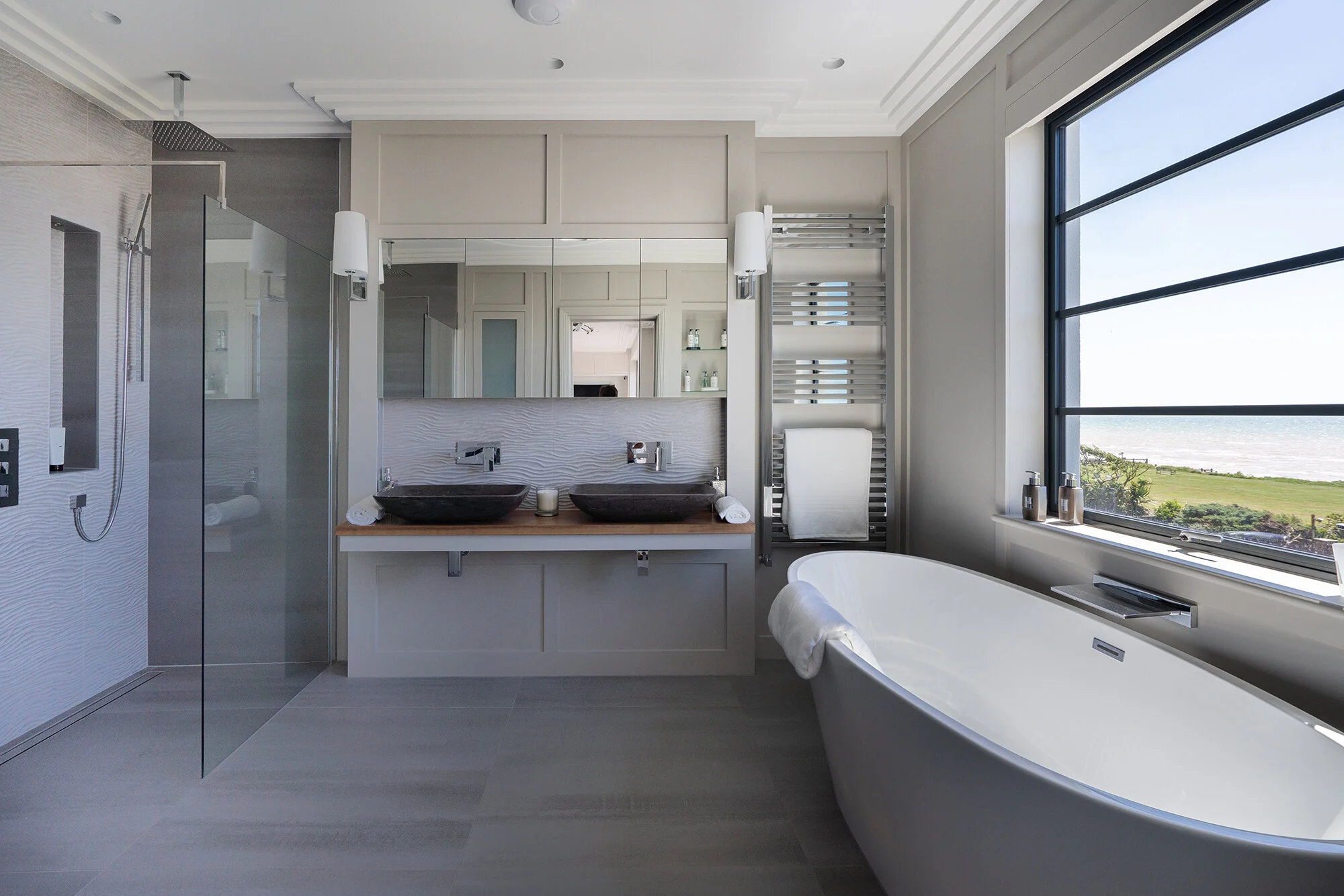 Modern bathroom with large freestanding bathtub next to a window, double sink vanity with black vessel sinks, wall-mounted faucets, a walk-in shower with a rain shower head, and a heated towel rail. The room has a coastal view through the large windo