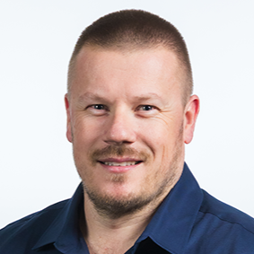 Smiling man with short hair and a beard, wearing a tan shirt, sitting in a room with shelves and a blue wall in the background.