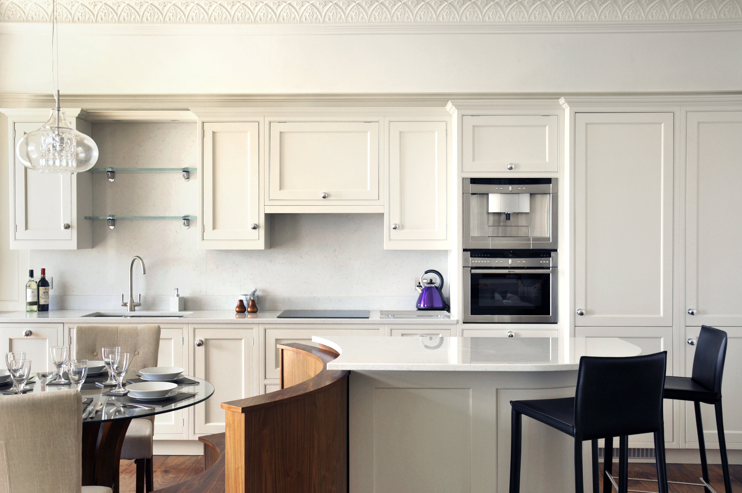 Modern kitchen with white cabinets, a black bar stool, glass dining table, and built-in appliances.