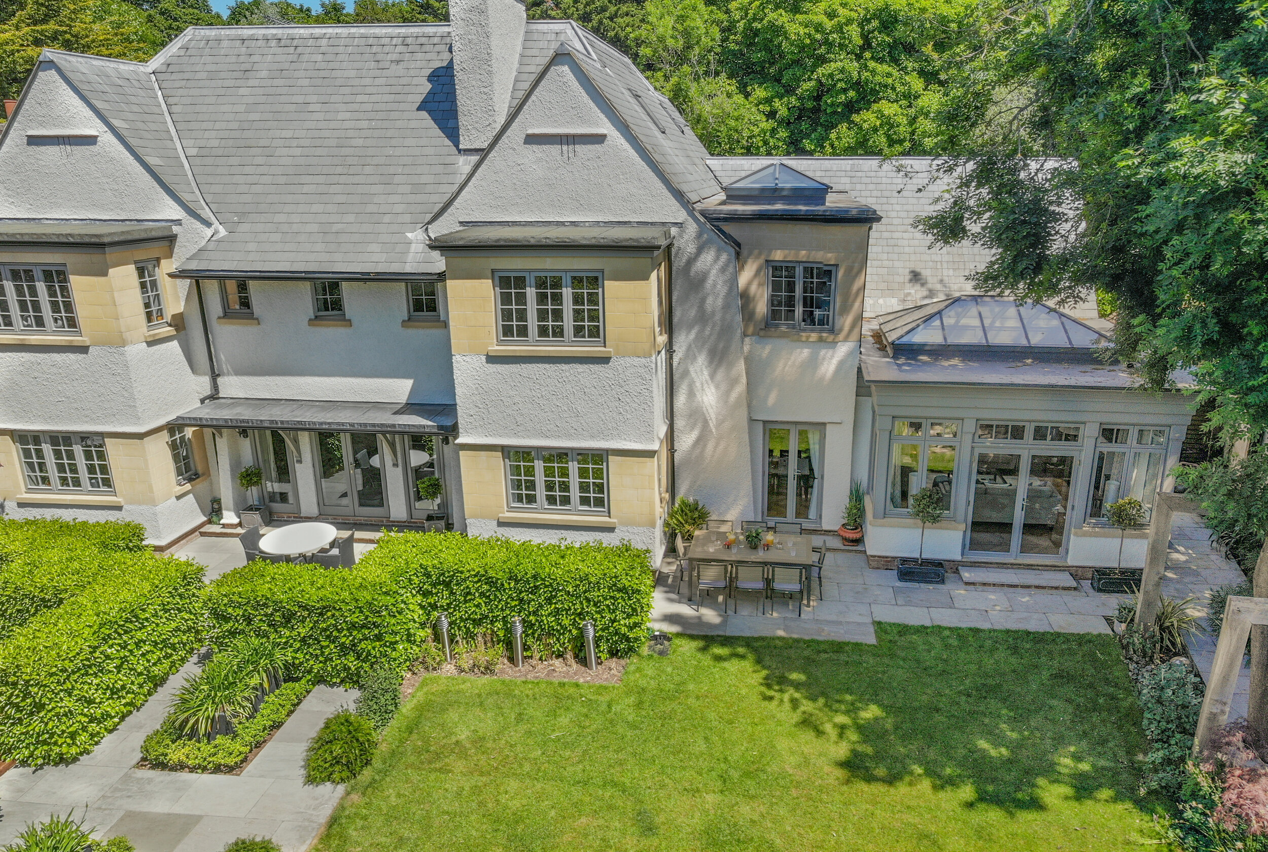 Aerial view of a large two-story house with gray roof and white exterior, featuring multiple windows and a sunroom. There is a well-maintained backyard with green lawn, shrubs, patio, and outdoor dining table.