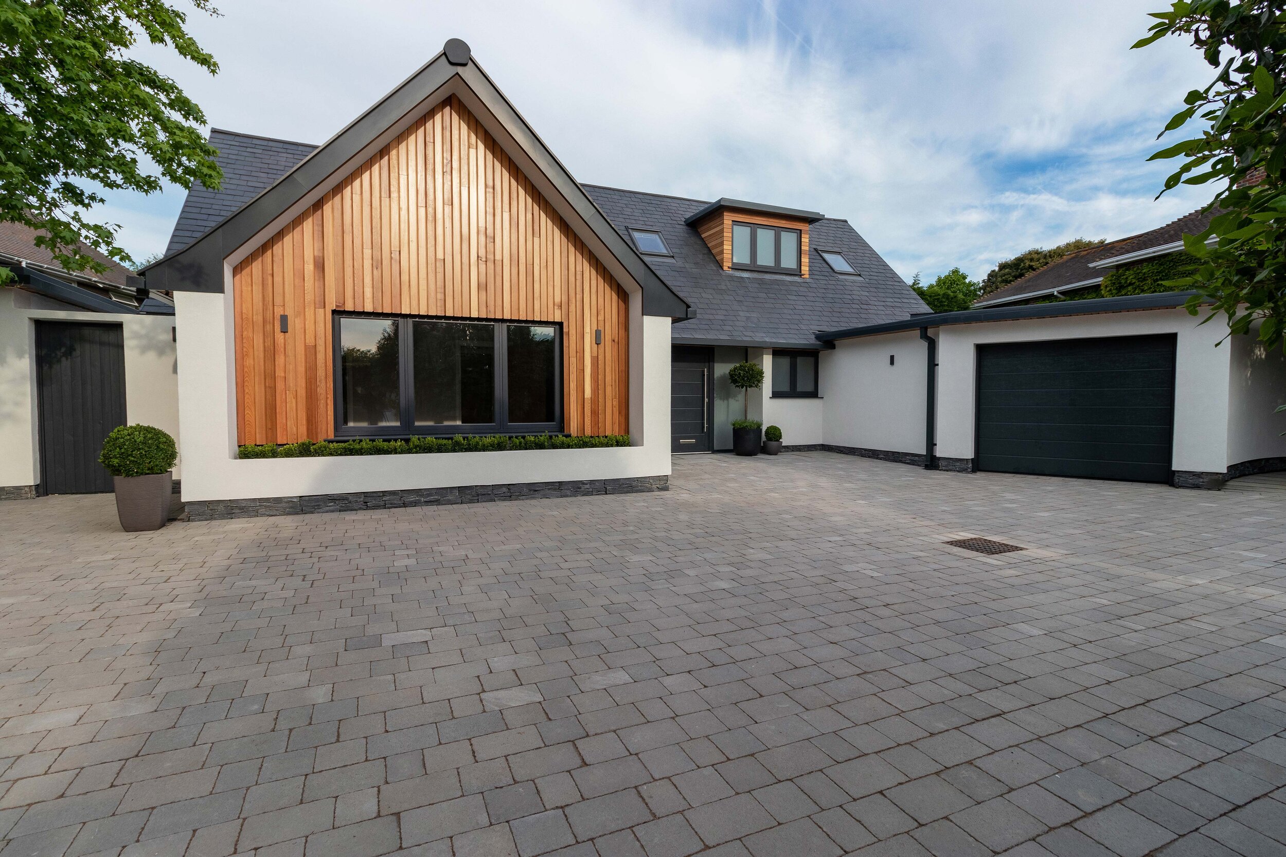 Modern single-story house with wood paneling, large windows, and a gray slate roof. Features a wide driveway with pavers, black garage door, and minimalistic landscaping.