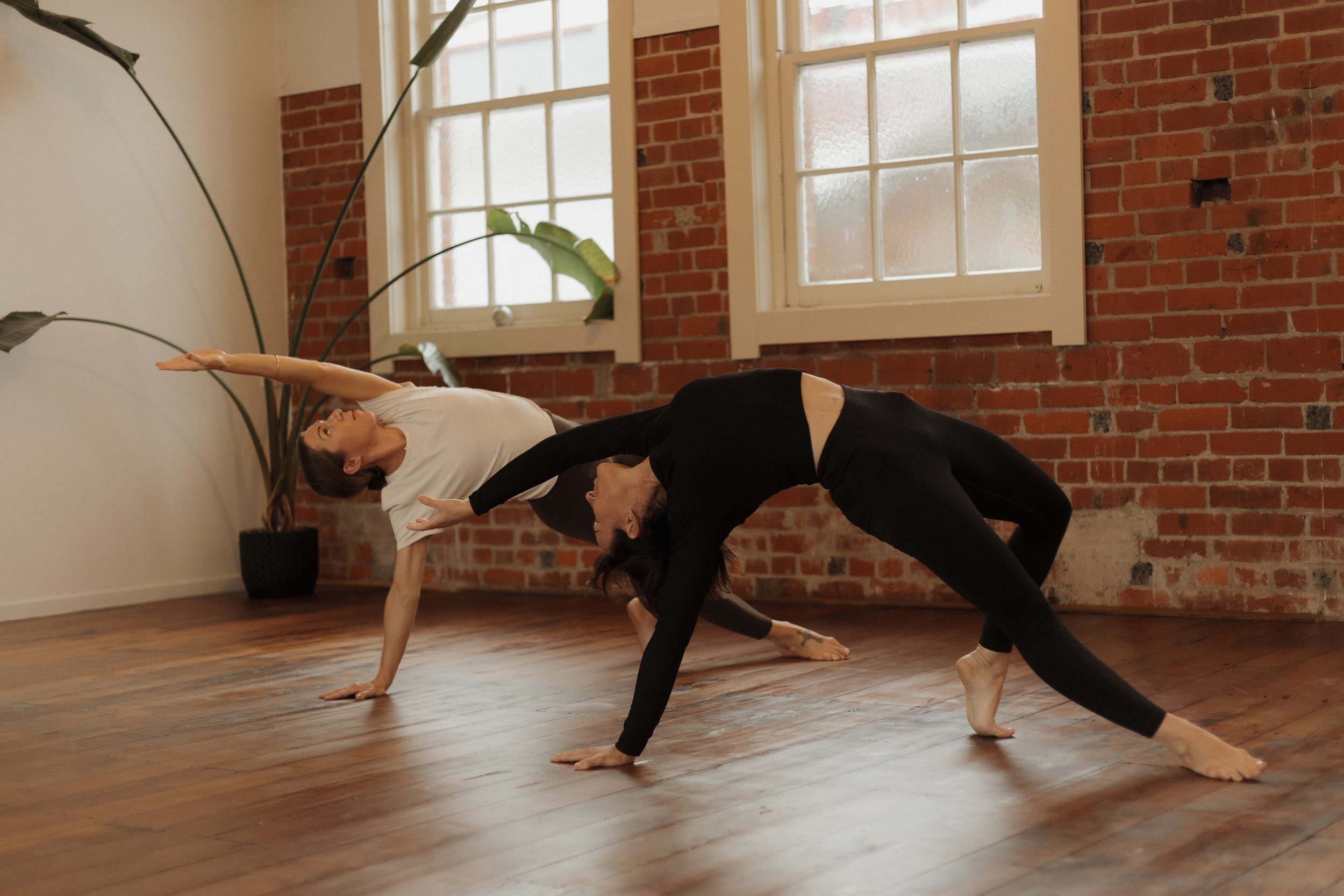 Two women practicing yoga in a studio with brick walls and large windows, one in a side plank pose and the other reaching towards her with one arm extended.