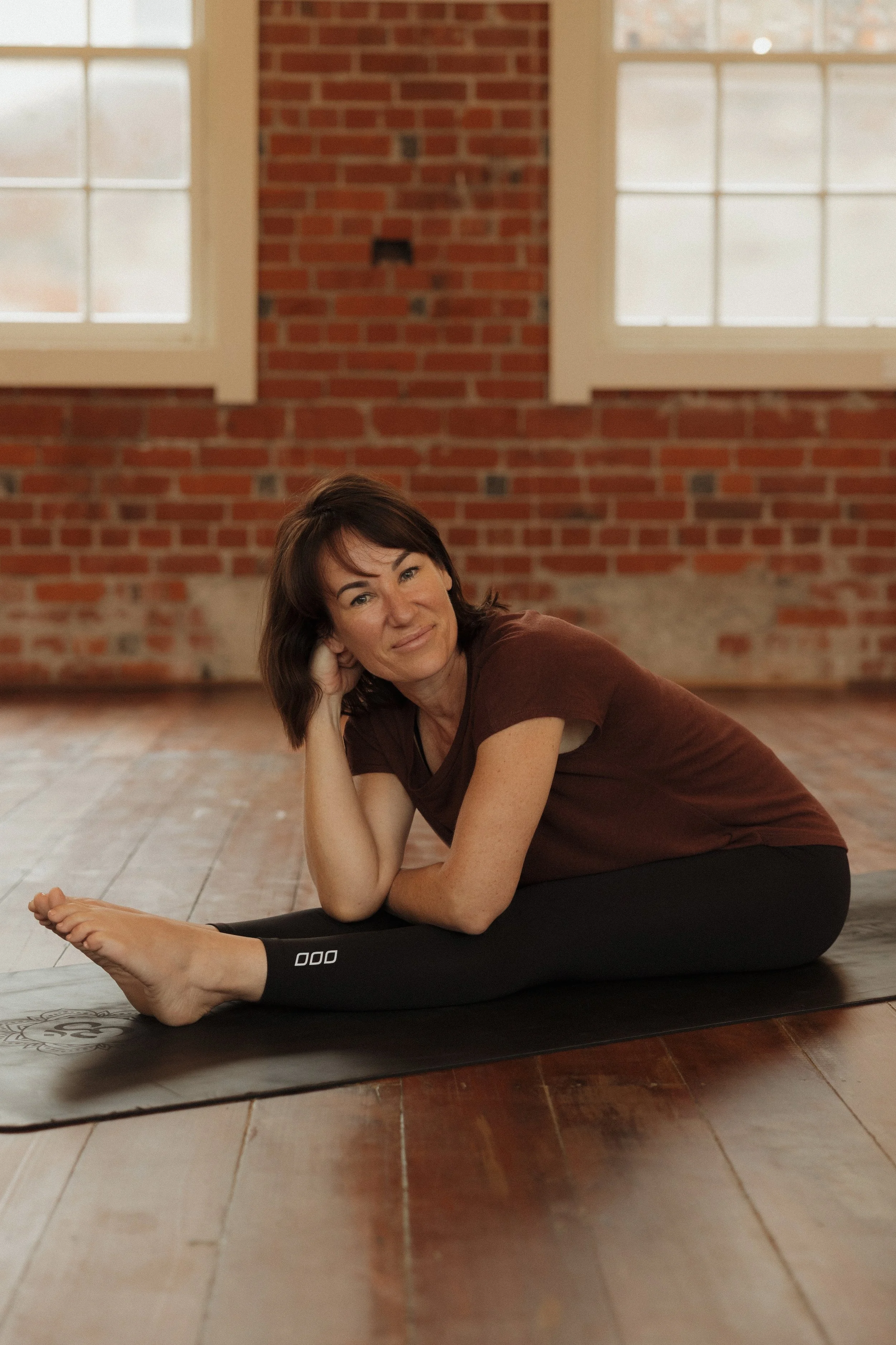 A woman with dark brown hair lying on a yoga mat in a brick-walled room with large windows, performing a seated forward bend stretch in a yoga or exercise session.
