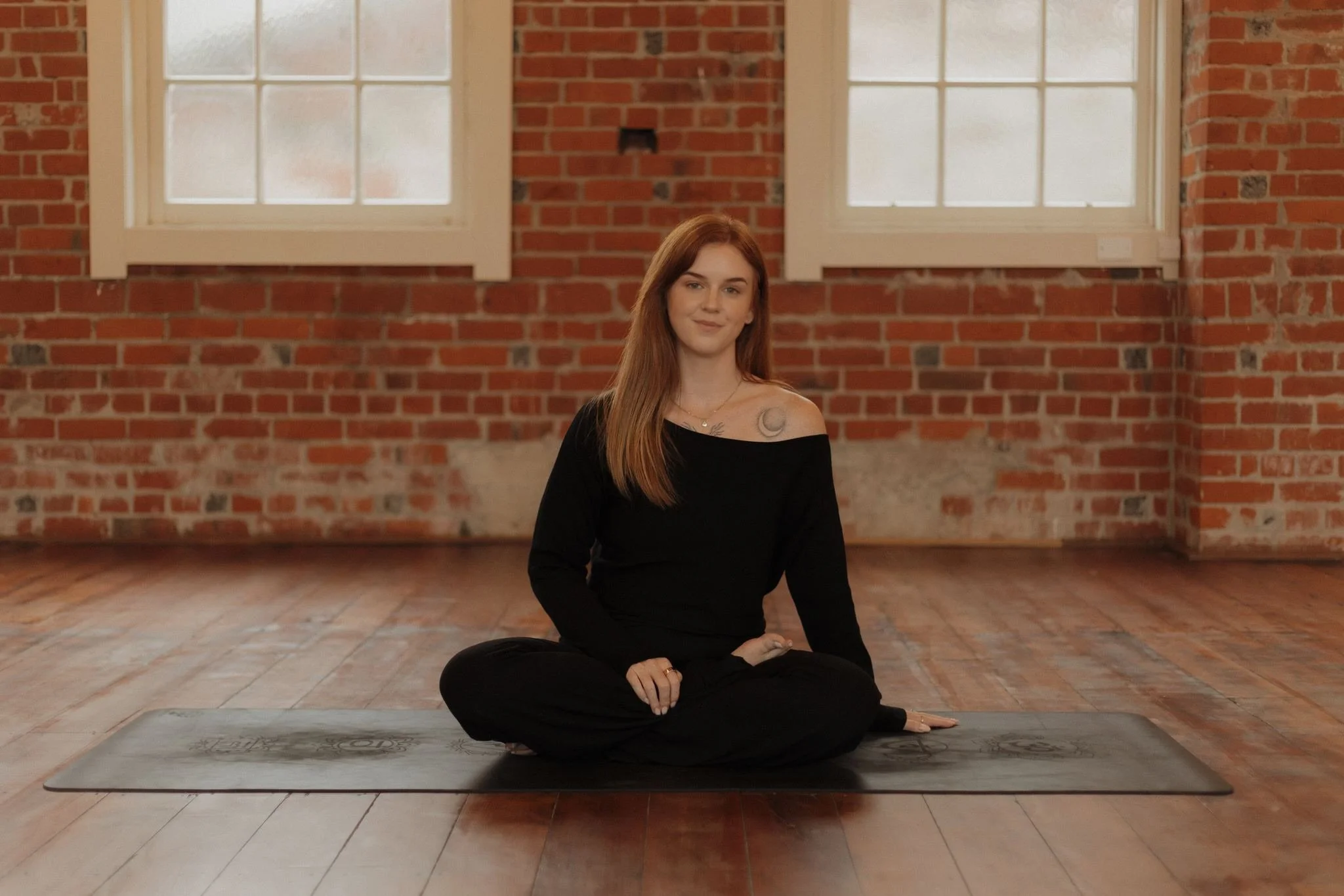 A woman with long red hair sitting cross-legged on a yoga mat in a room with brick walls and large windows, practicing yoga or meditative postures.