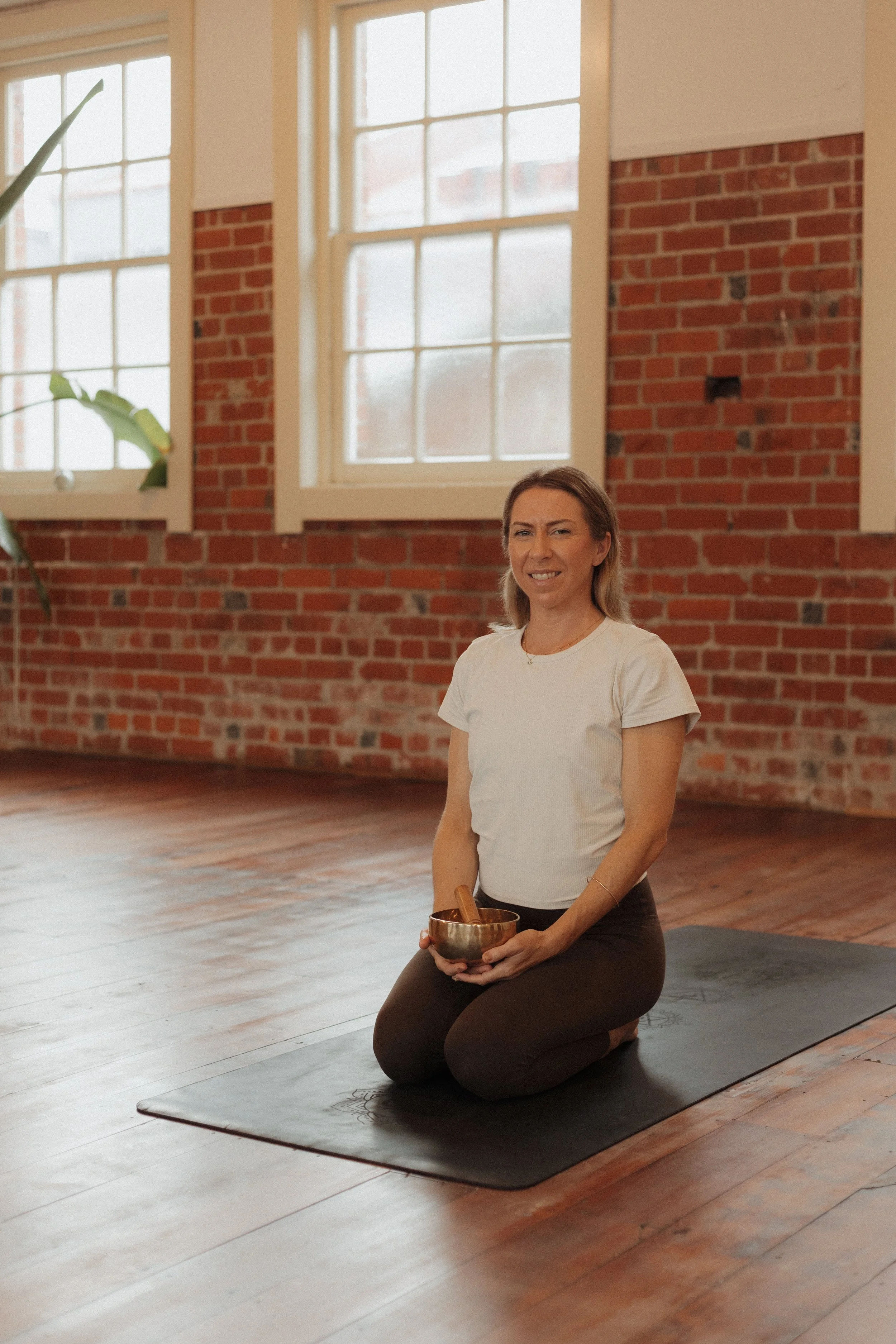 A woman kneeling on a yoga mat in a bright room with brick walls and large windows, holding a singing bowl with a wooden mallet.