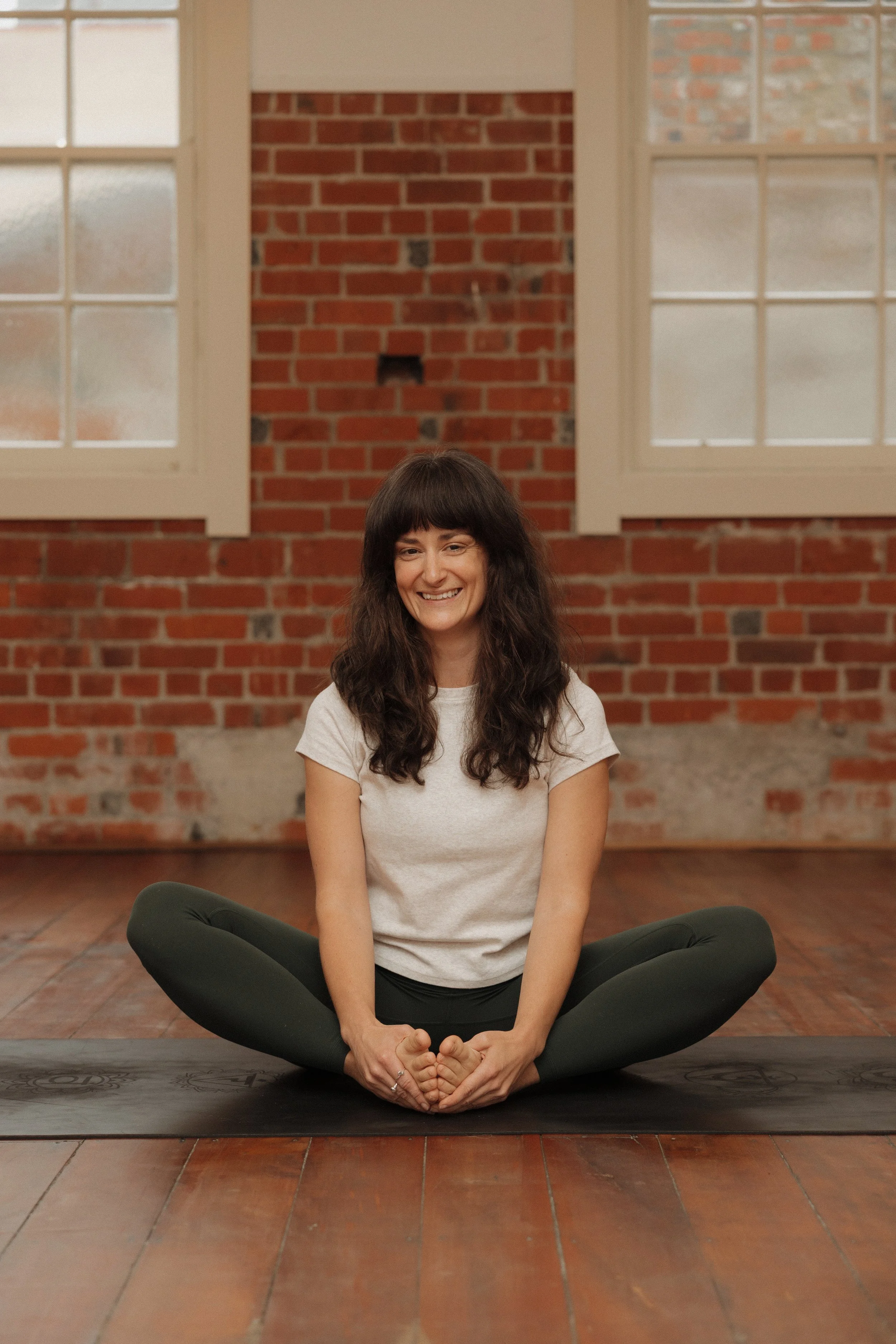 A woman with long brown hair sitting cross-legged on a yoga mat in a brick-walled room, smiling.