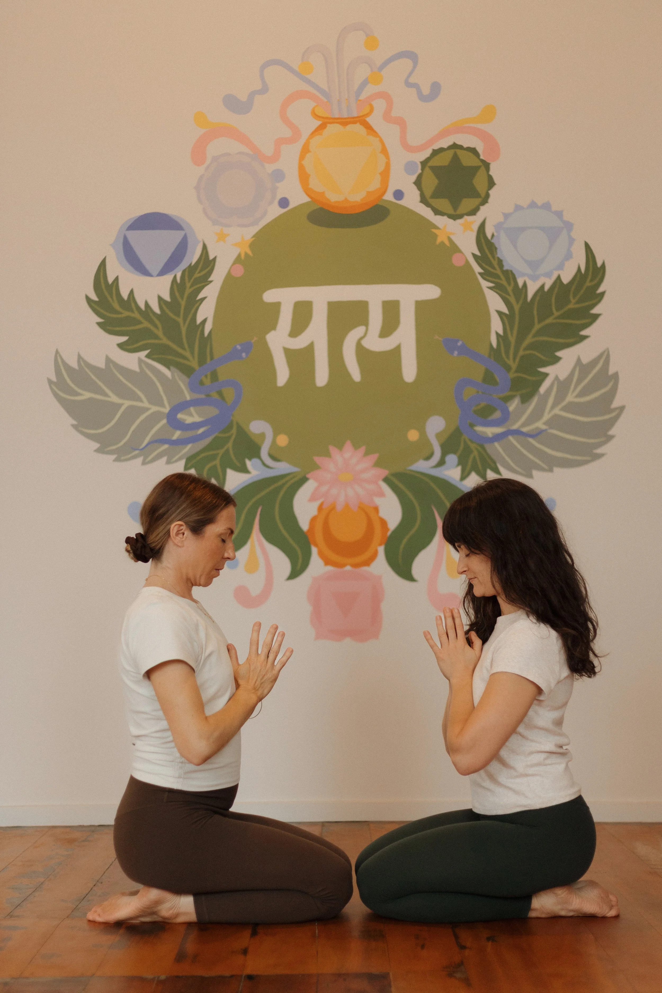 Two women practicing yoga in a studio, kneeling on yoga mats with hands pressed together in a prayer position, facing each other, with a colorful decorative wall art featuring various shapes and a large green circle with jointed fingers in the background.
