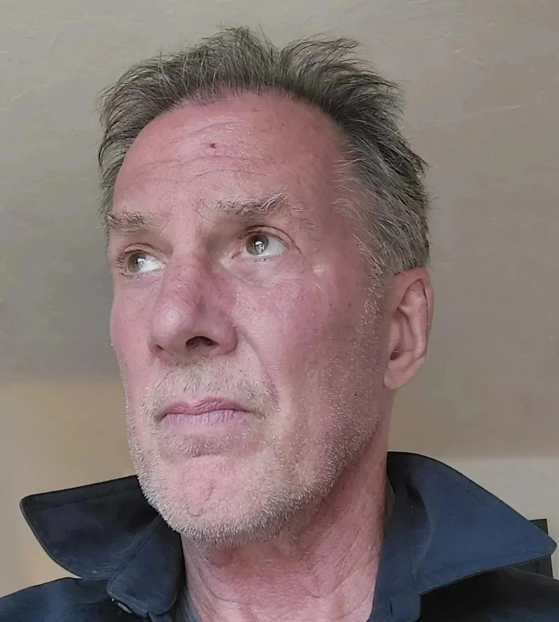 A close-up of an older man with gray hair, a beard, and blue eyes, wearing a dark jacket with the collar up, looking upwards and to the left.