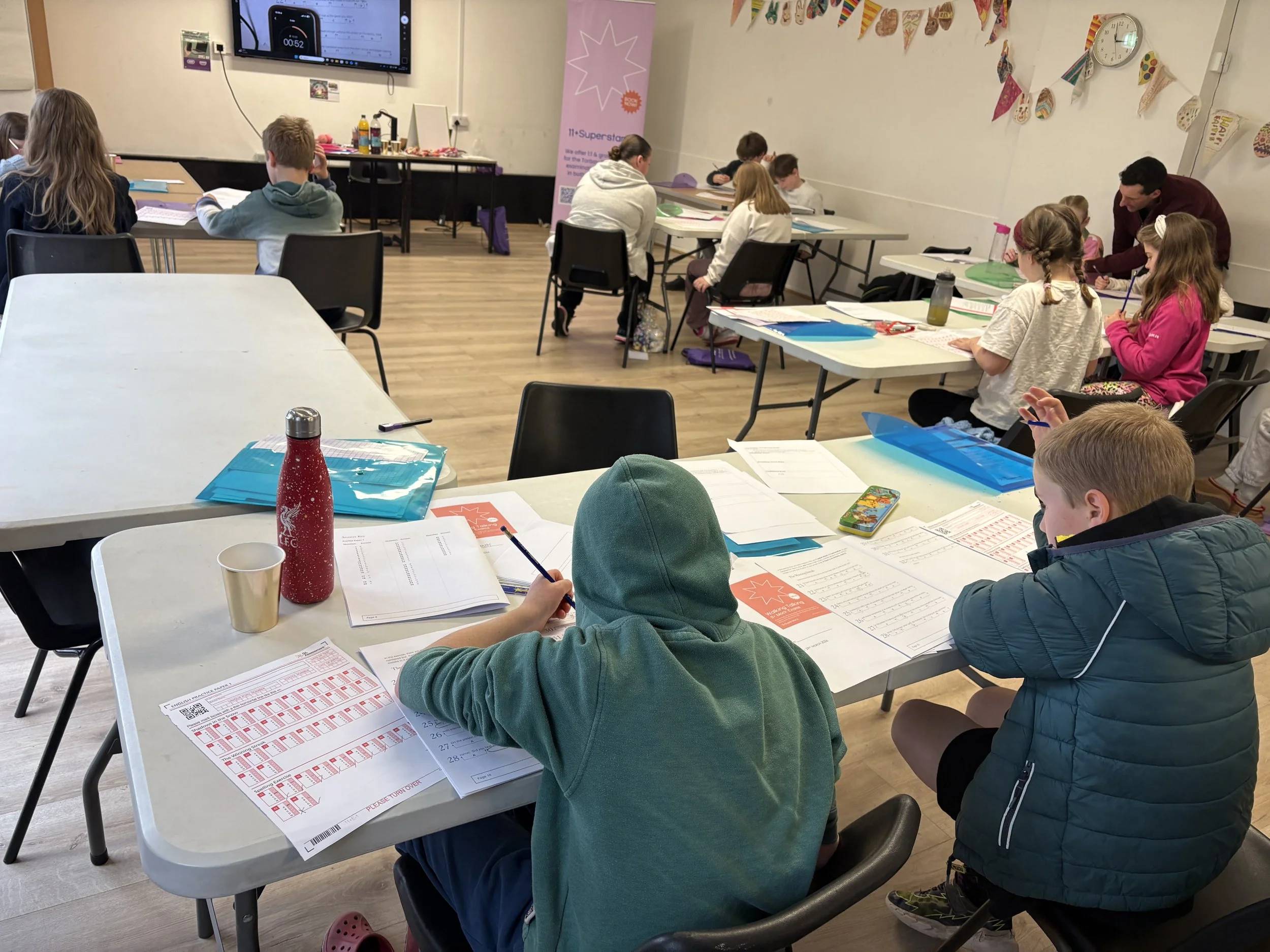 Children and adults sitting at tables in a classroom, working on papers, with a large screen on the wall, hanging decorations, and a clock on the wall.