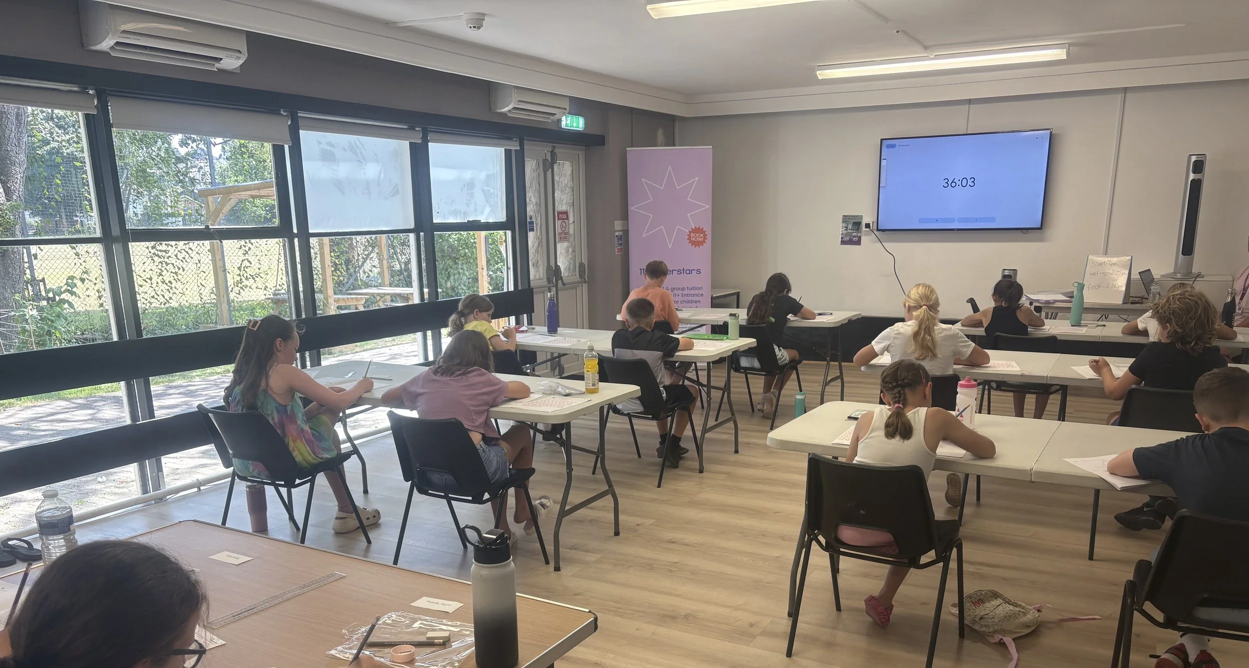 Children sitting at desks in a classroom listening to a teacher, with a large screen displaying a countdown timer.