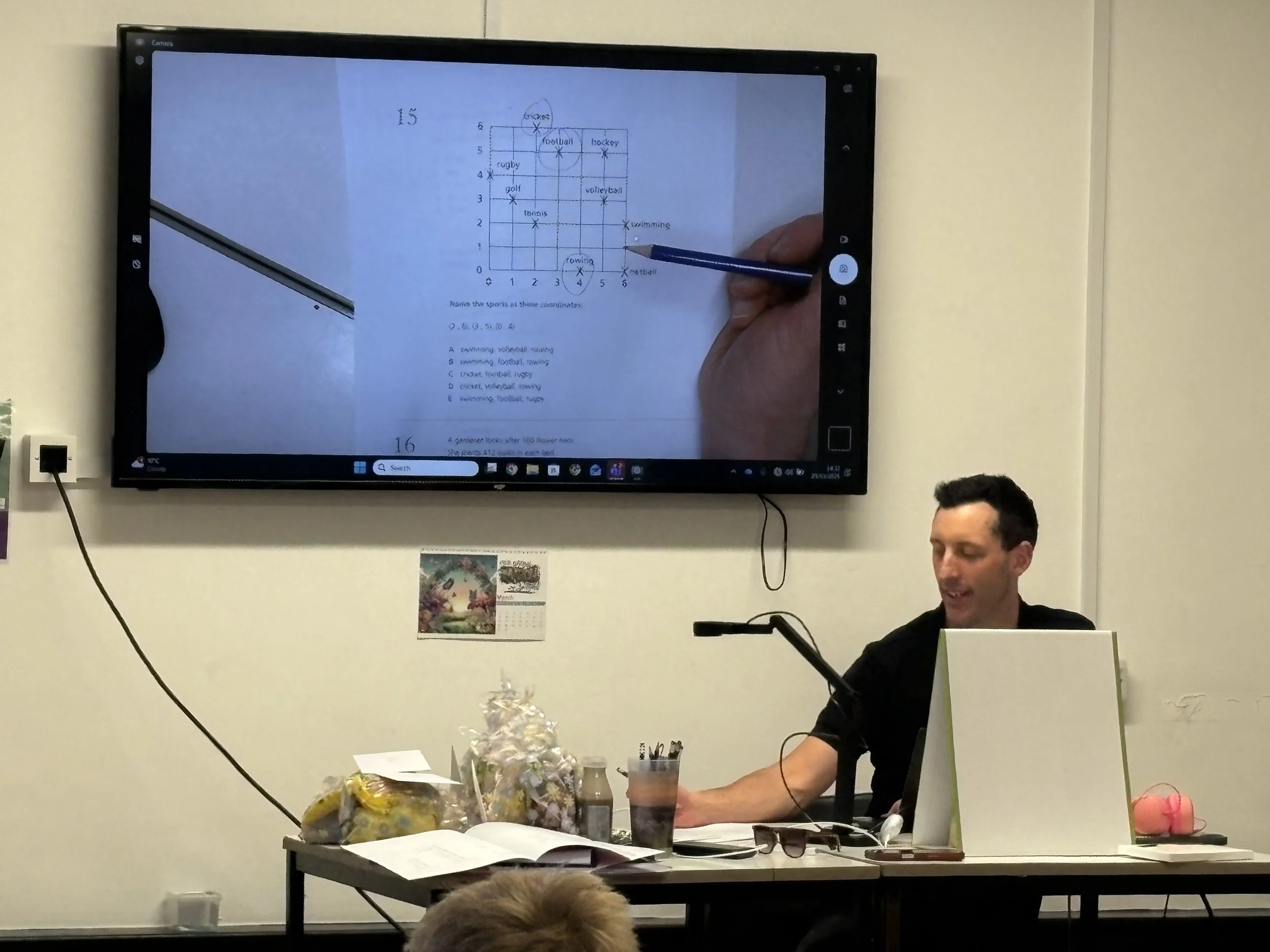 A person with dark hair and glasses is sitting at a table, looking at a monitor. Office supplies and snacks are on the table. A large screen behind him displays a handwritten sports schedule or chart with various sports names and numbers.