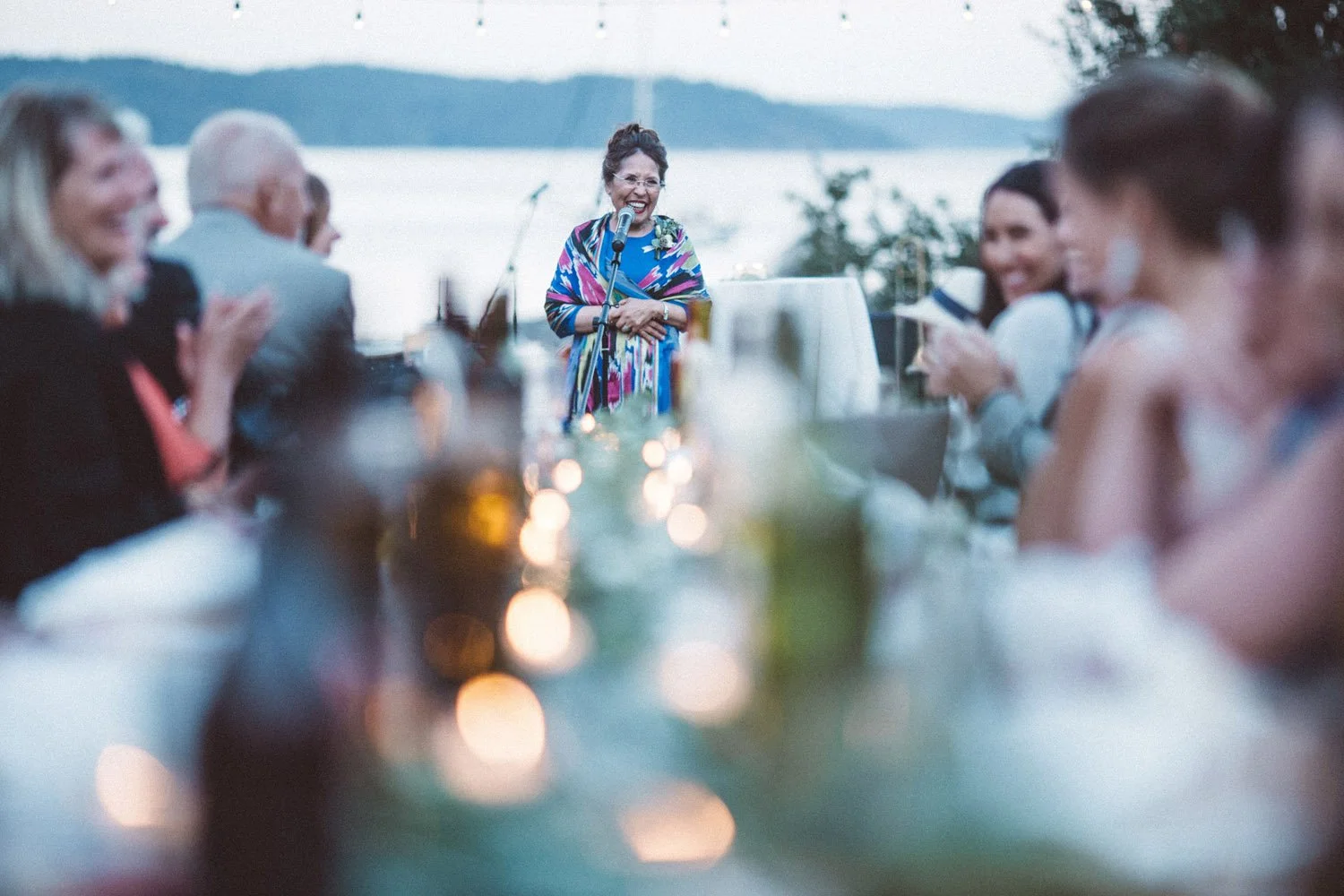 A long dining table outside at dusk at Pebble Cove Farm, with wedding guest listening to a speech and lots of small lit tea lights in the center of the table.