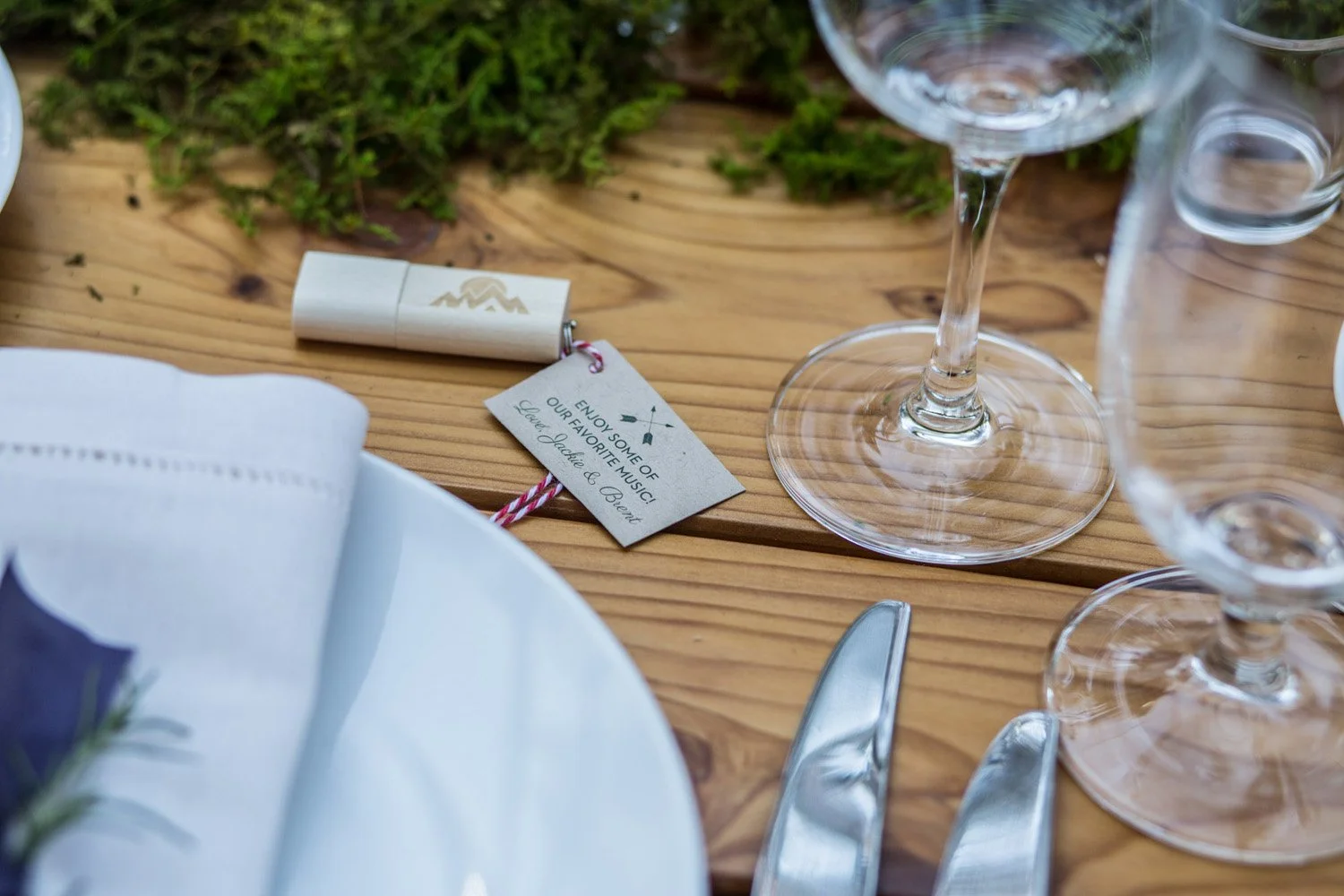 A close up view of a wooden memory stick, laid next to a plate setting on a wooden table. It has a small label on it saying 'Listen to some of our favorite music'.