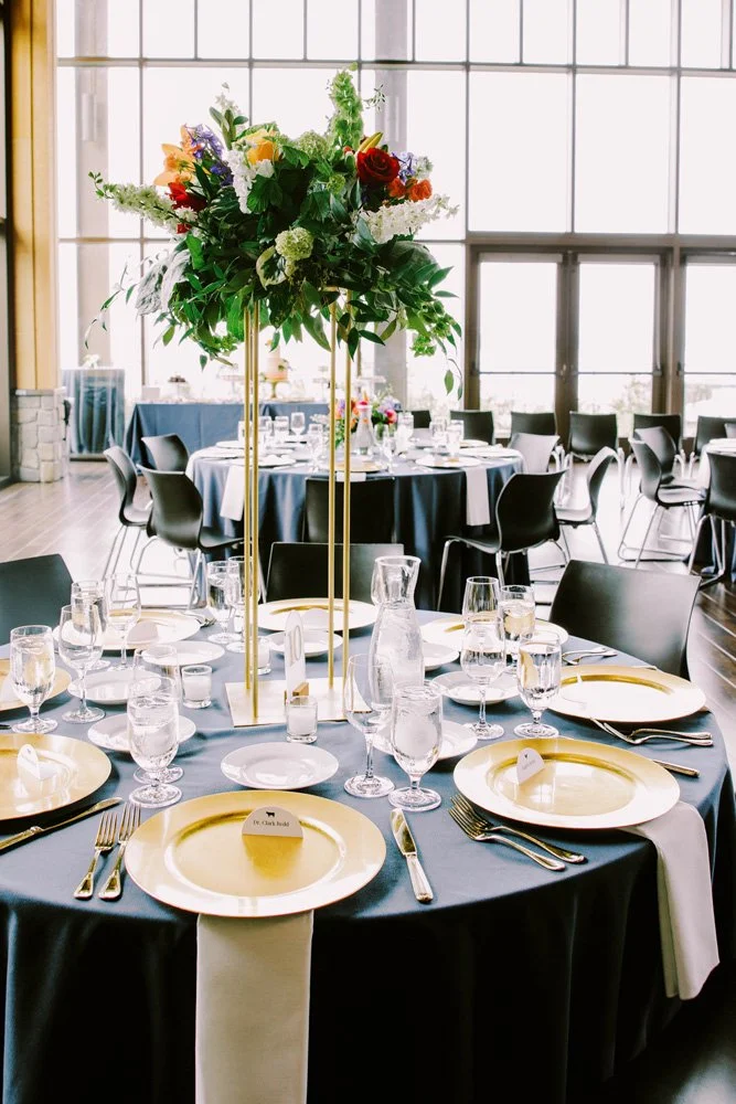 Round tables with navy table linens, gold plates and tall vibrant colored floral centerpieces Aviation boarding pass inspired seating place cards at the Rosehill Center-Photography by Jenn Tai & Co.-planned and designed by Simply Wed