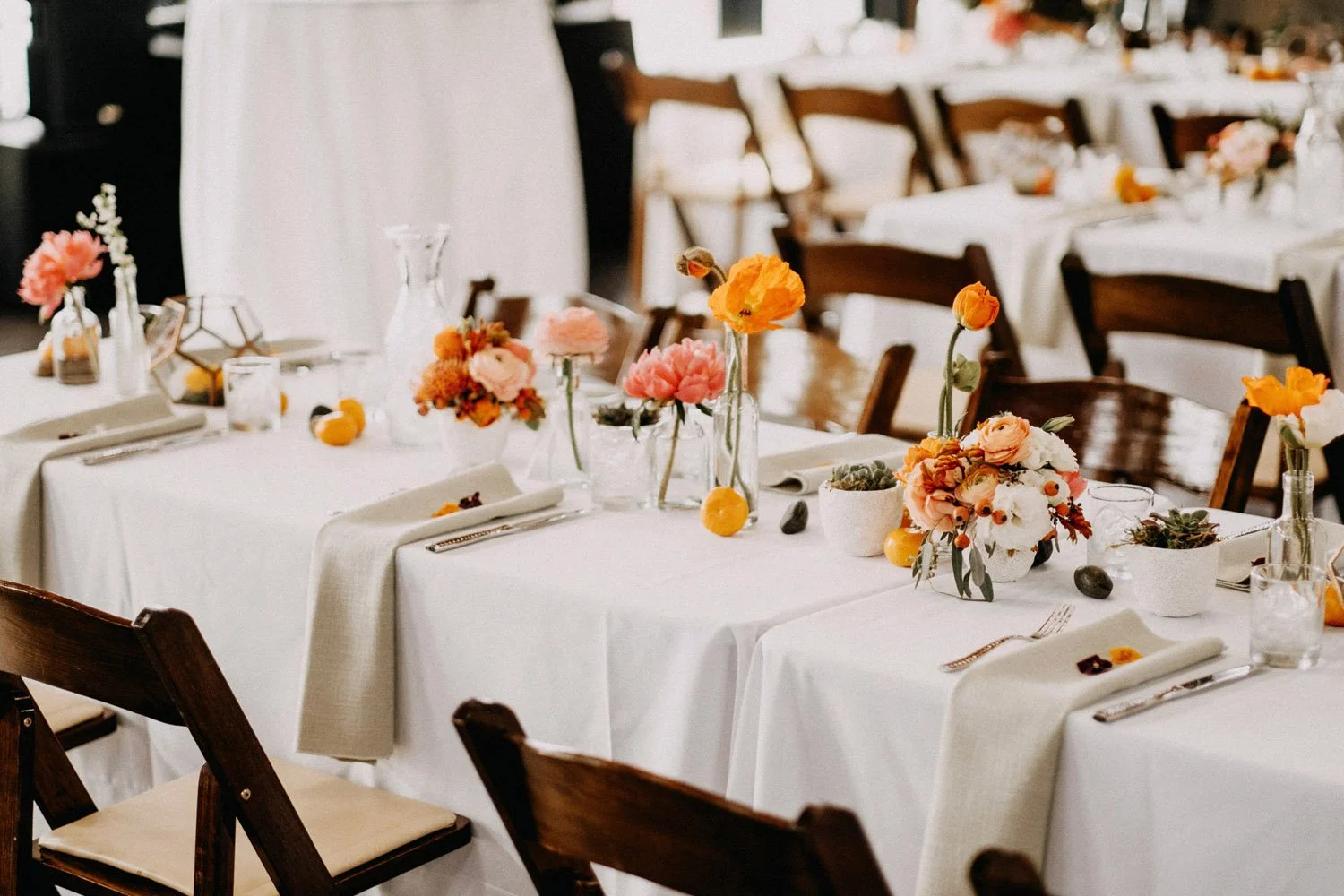 Warm wood toned farmhouse tables with crisp white table linens with pops of vibrant orange and coral peonies in small vases and clementines mixed in at the stunning Golden Gardens Bath House-Photography by Dylan M Howell Photography-planned and desig