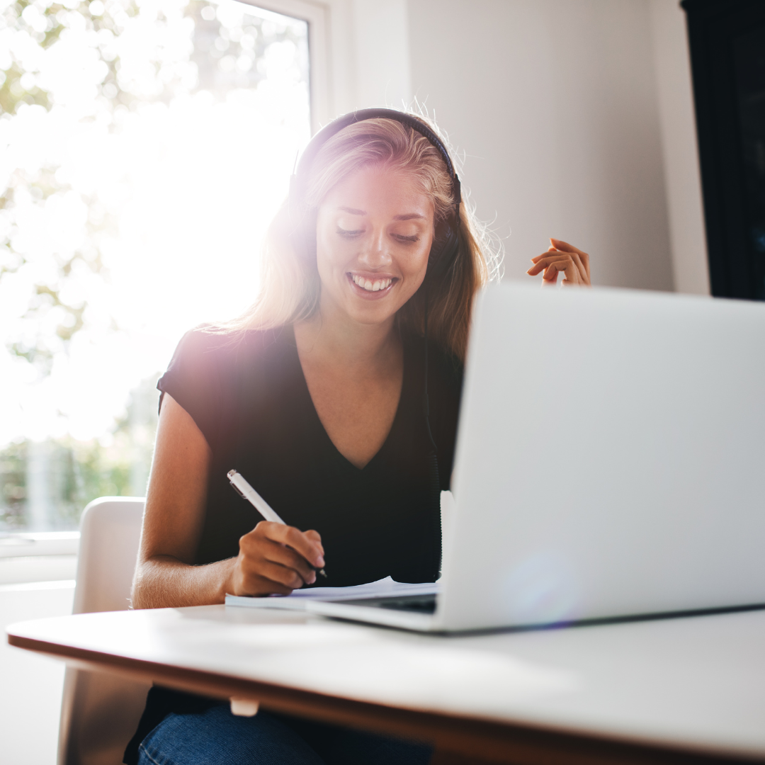 Woman studying remotely. Learning english happily wearing headphones in front of her laptop whilst taking English notes.