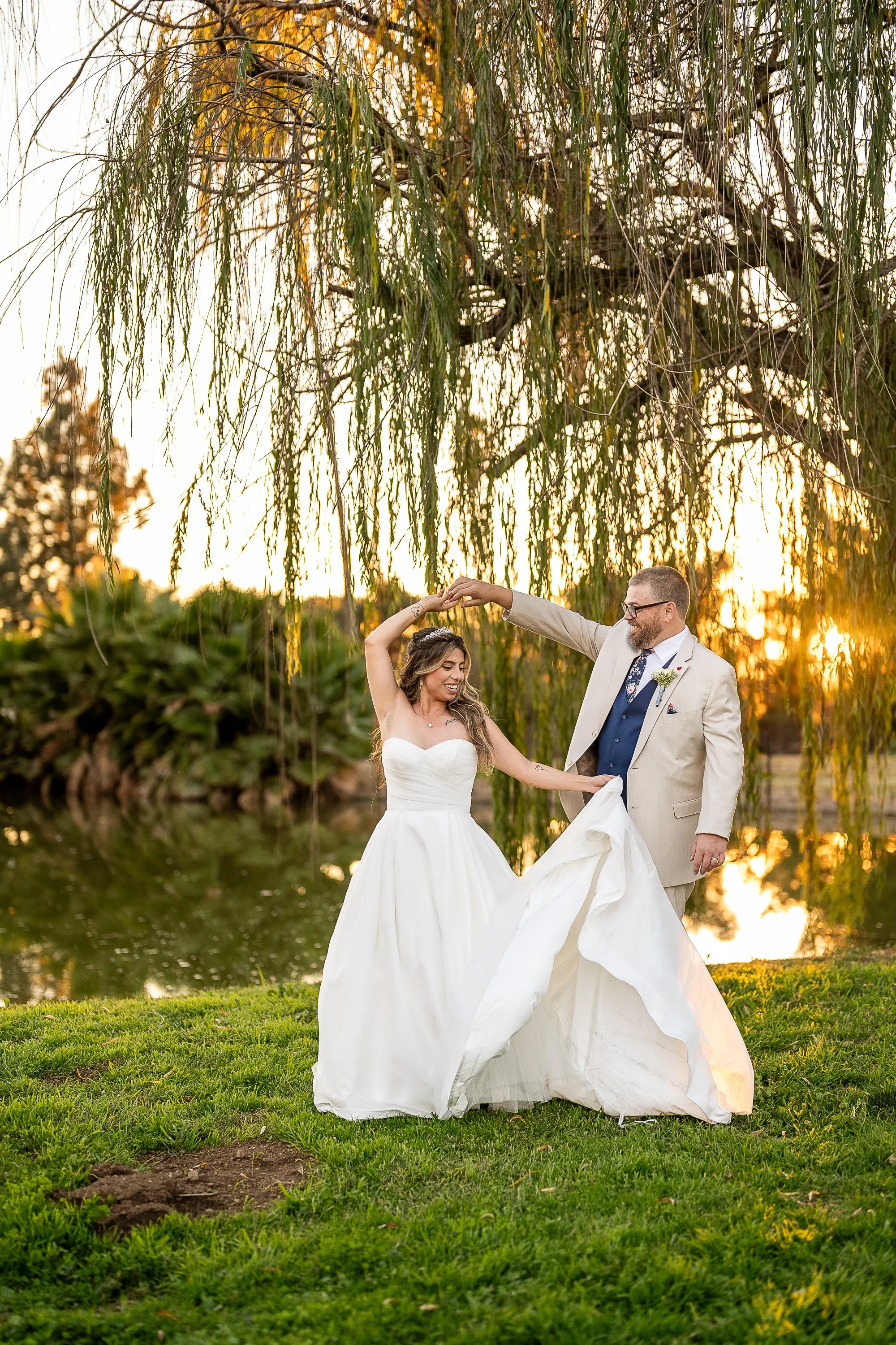 A bride and groom dancing outside near a pond at sunset, with the groom in a light-colored suit and the bride in a strapless white wedding dress.