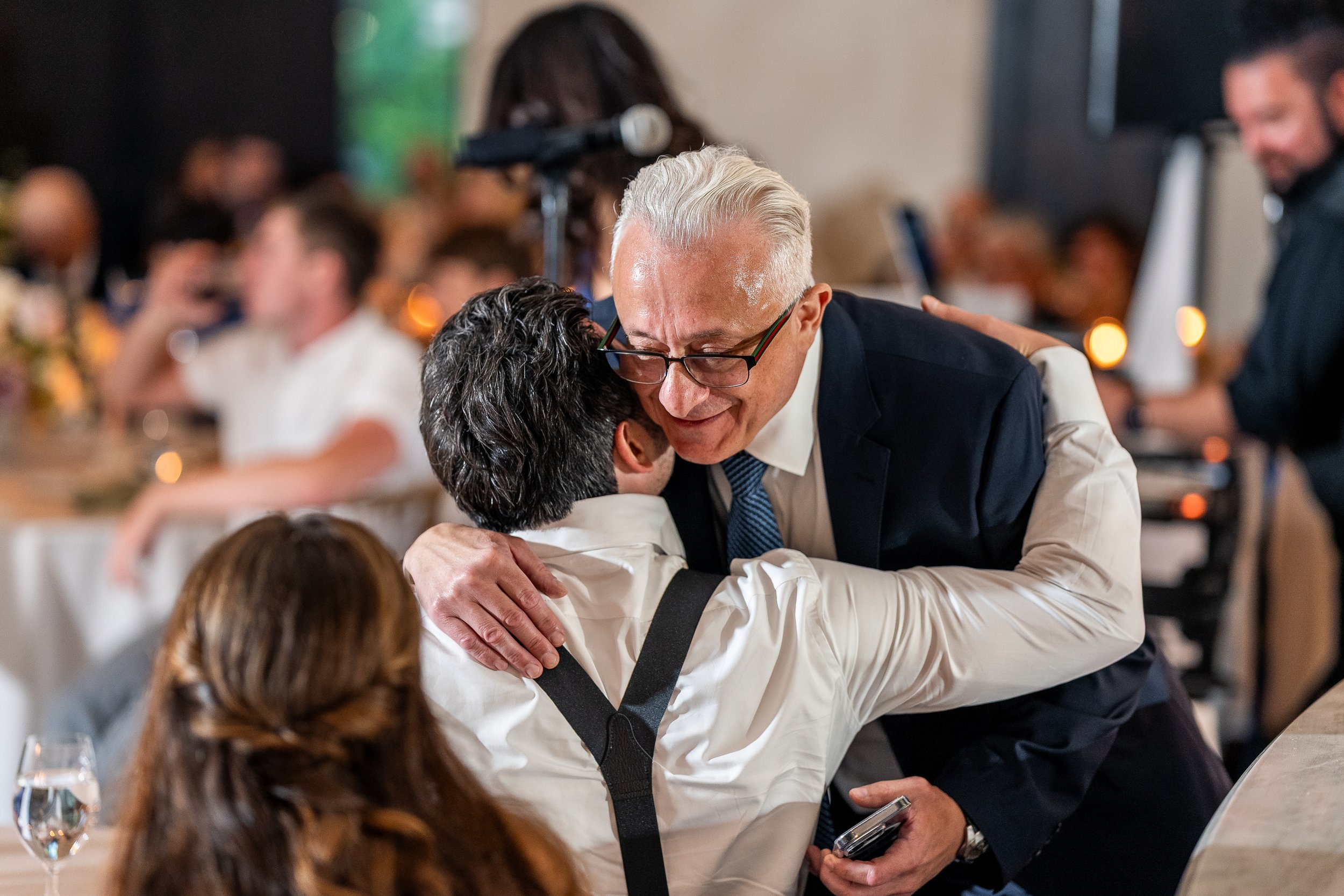 An older man with glasses and a suit hugging a younger man seated at a table, in a warmly lit gathering or celebration setting.